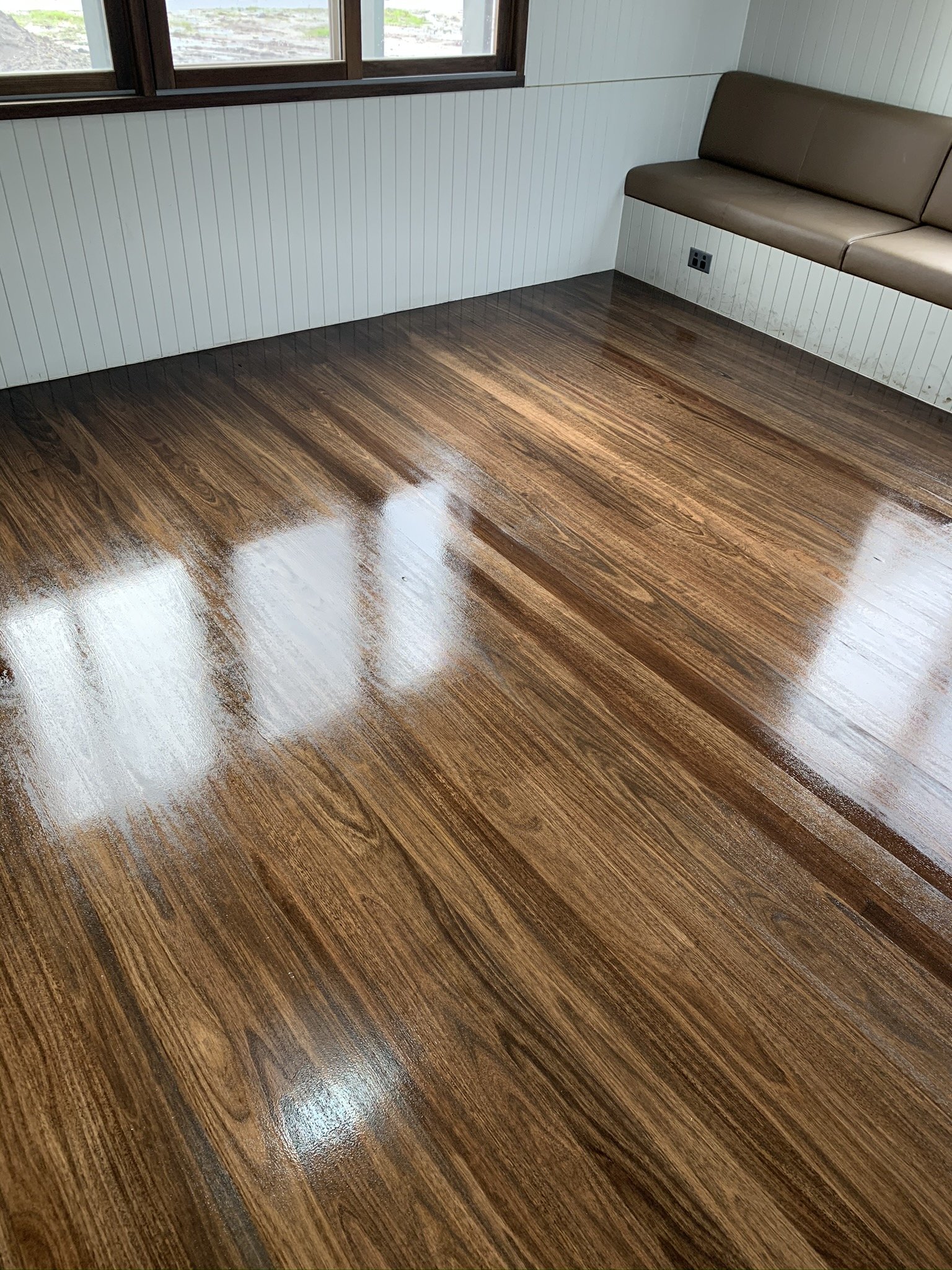 Shiny, polished hardwood floor in a room with white paneled walls and a brown bench along the wall. Window reflecting light on the floor.
