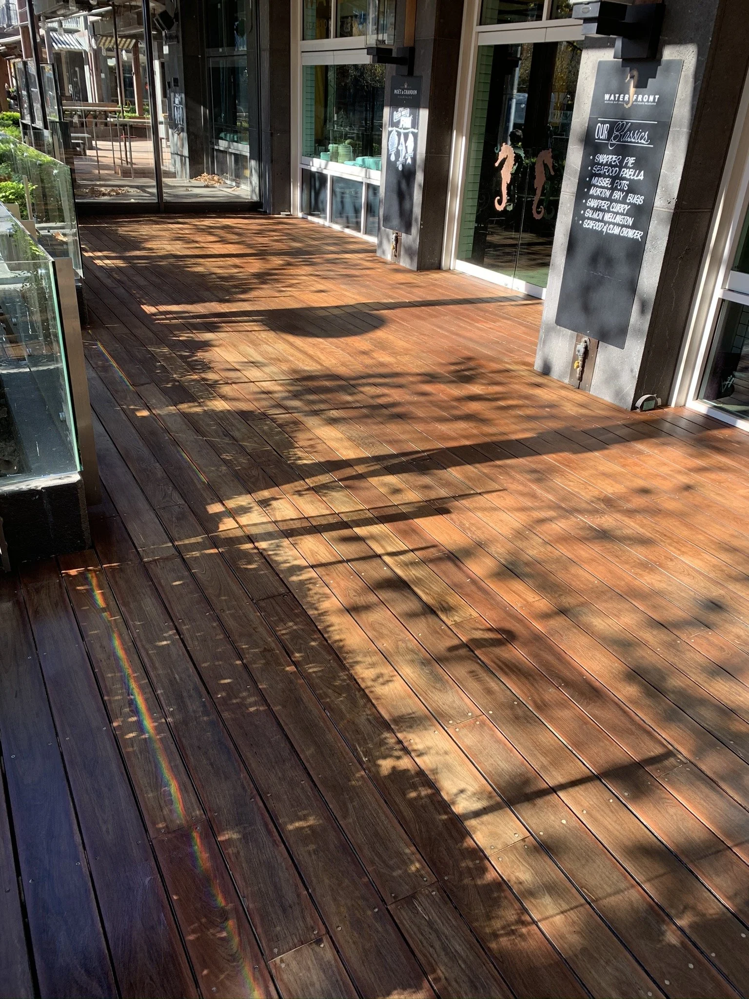 Wooden outdoor patio with shadows cast by nearby trees and a faint rainbow reflection on the floor. There are glass walls with a door and chalkboards listing menu items.