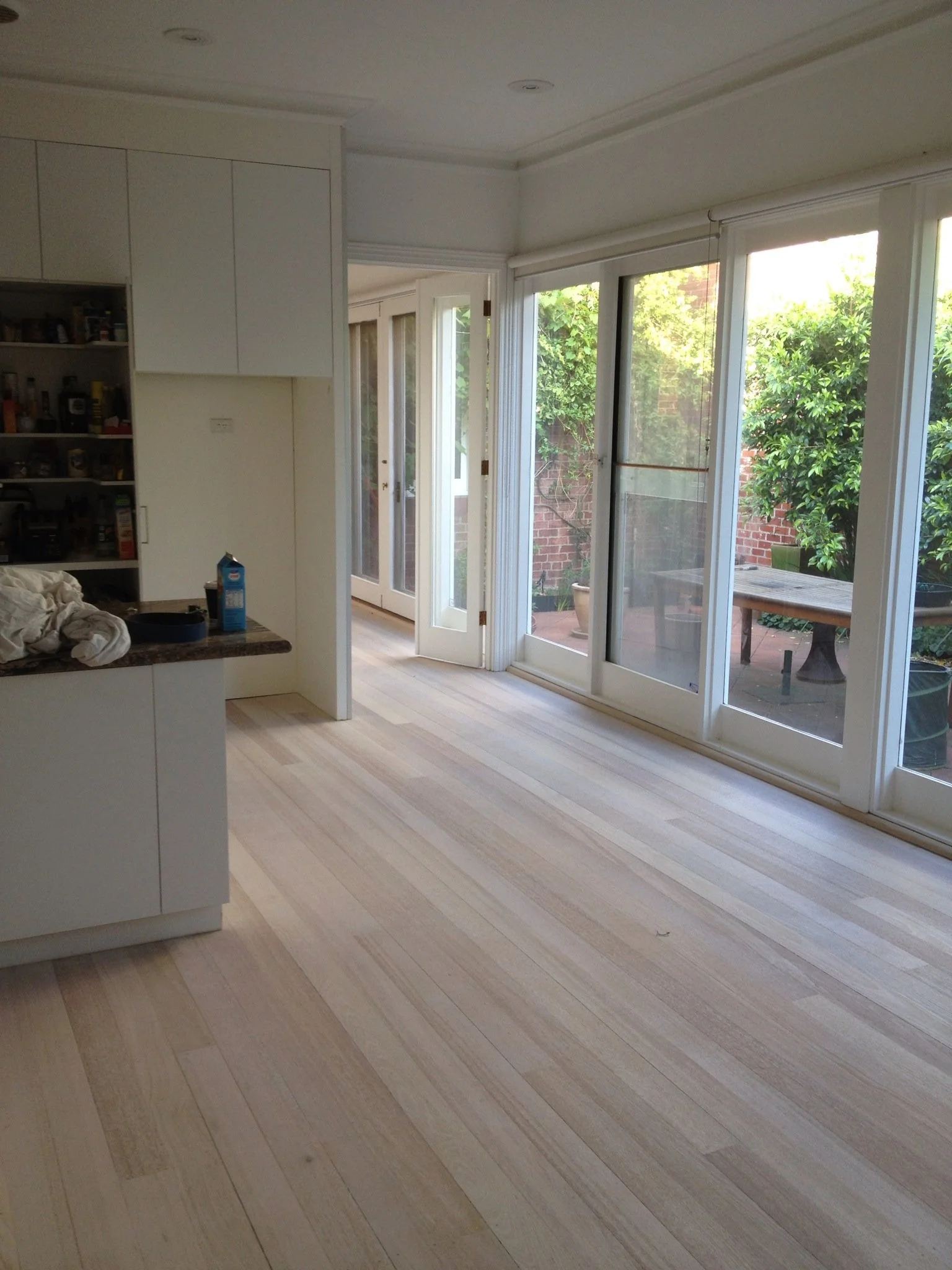 Empty kitchen with light wood flooring, white cabinets, and large sliding glass doors leading to a backyard with greenery and a patio table.