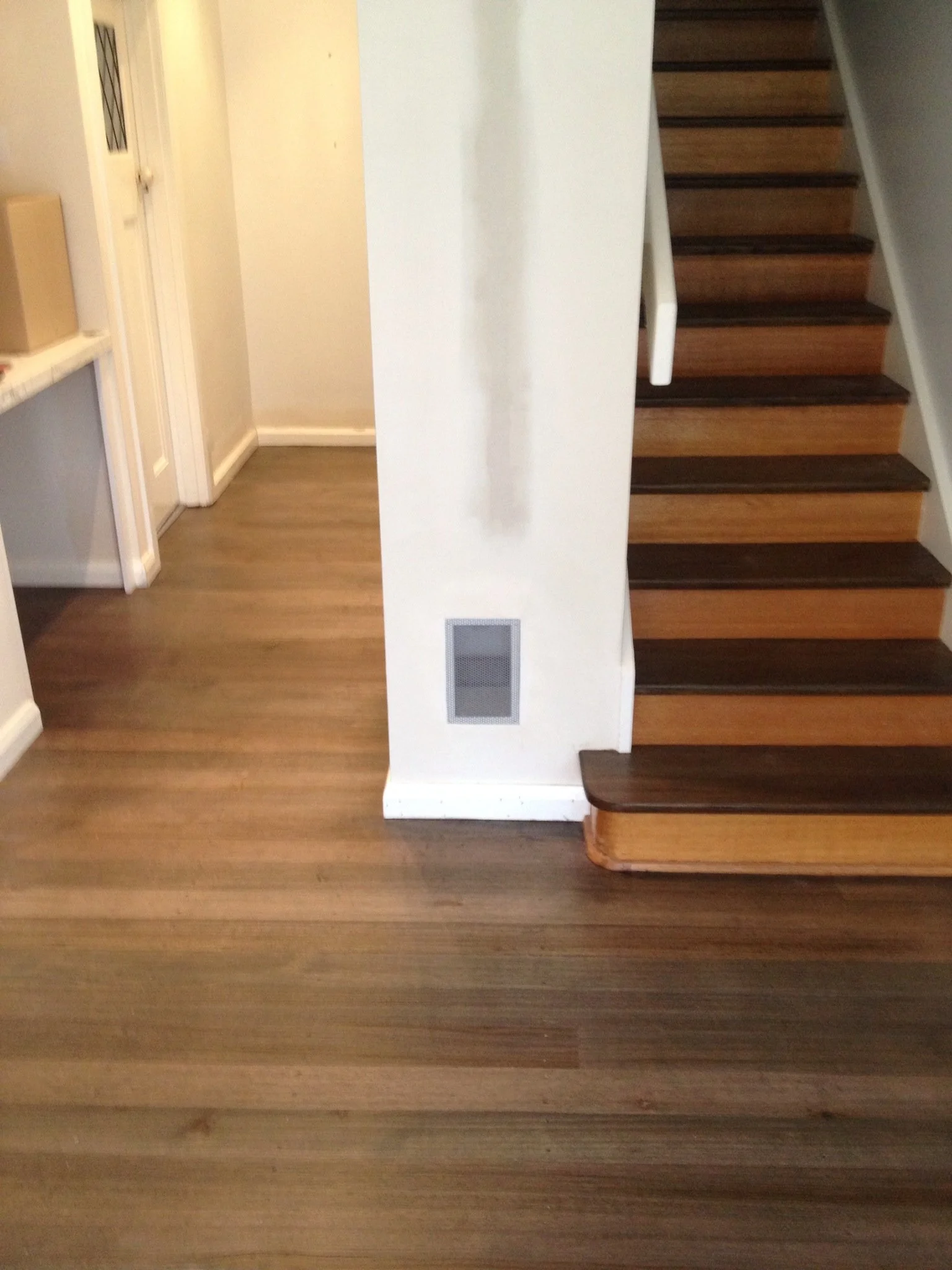Interior view of a home with wooden flooring, a staircase with dark wooden treads, and a white wall with a vent.