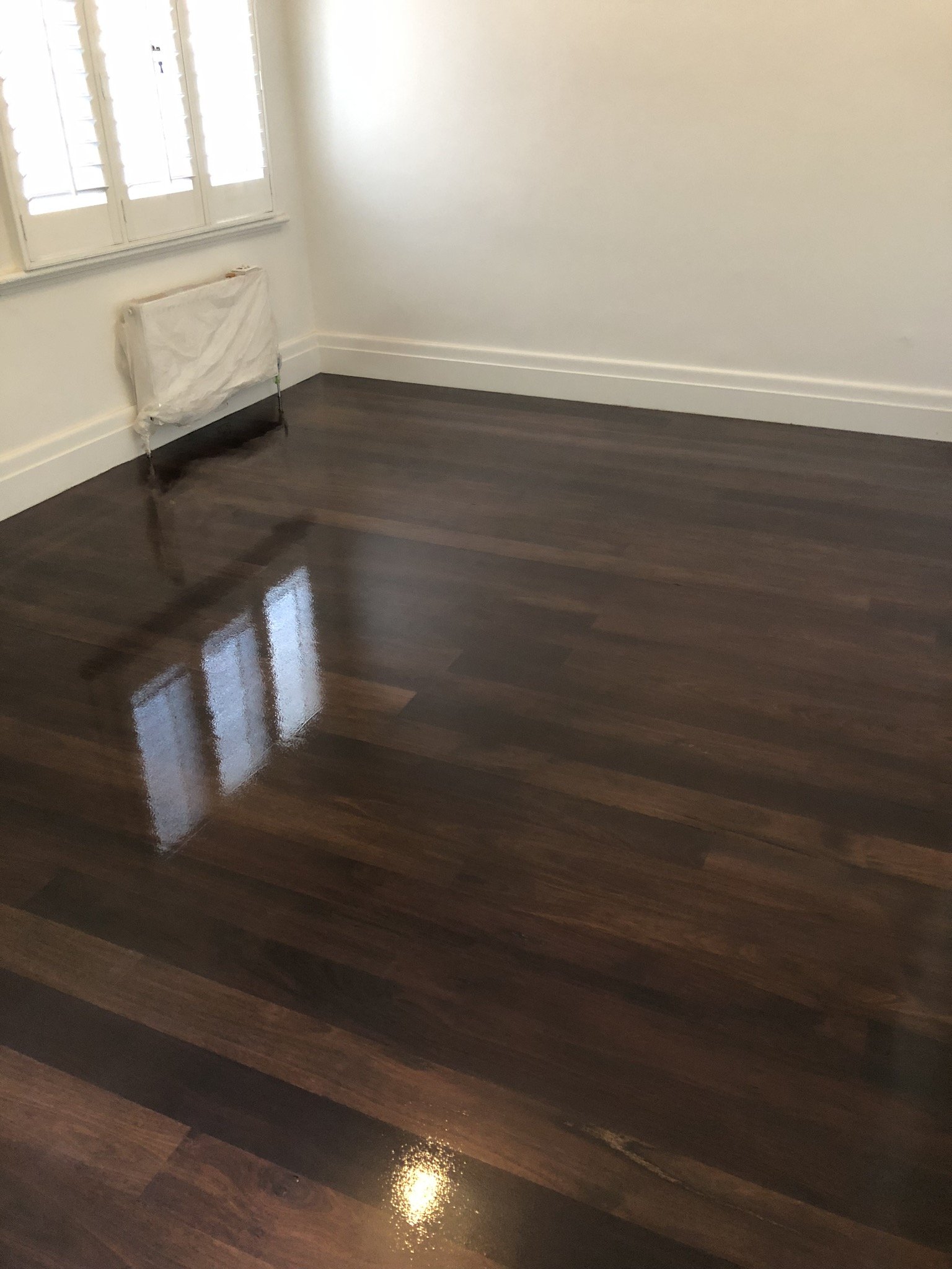 Empty room with dark polished wooden floor, white walls, window with shutters, and a small radiator covered with cloth.