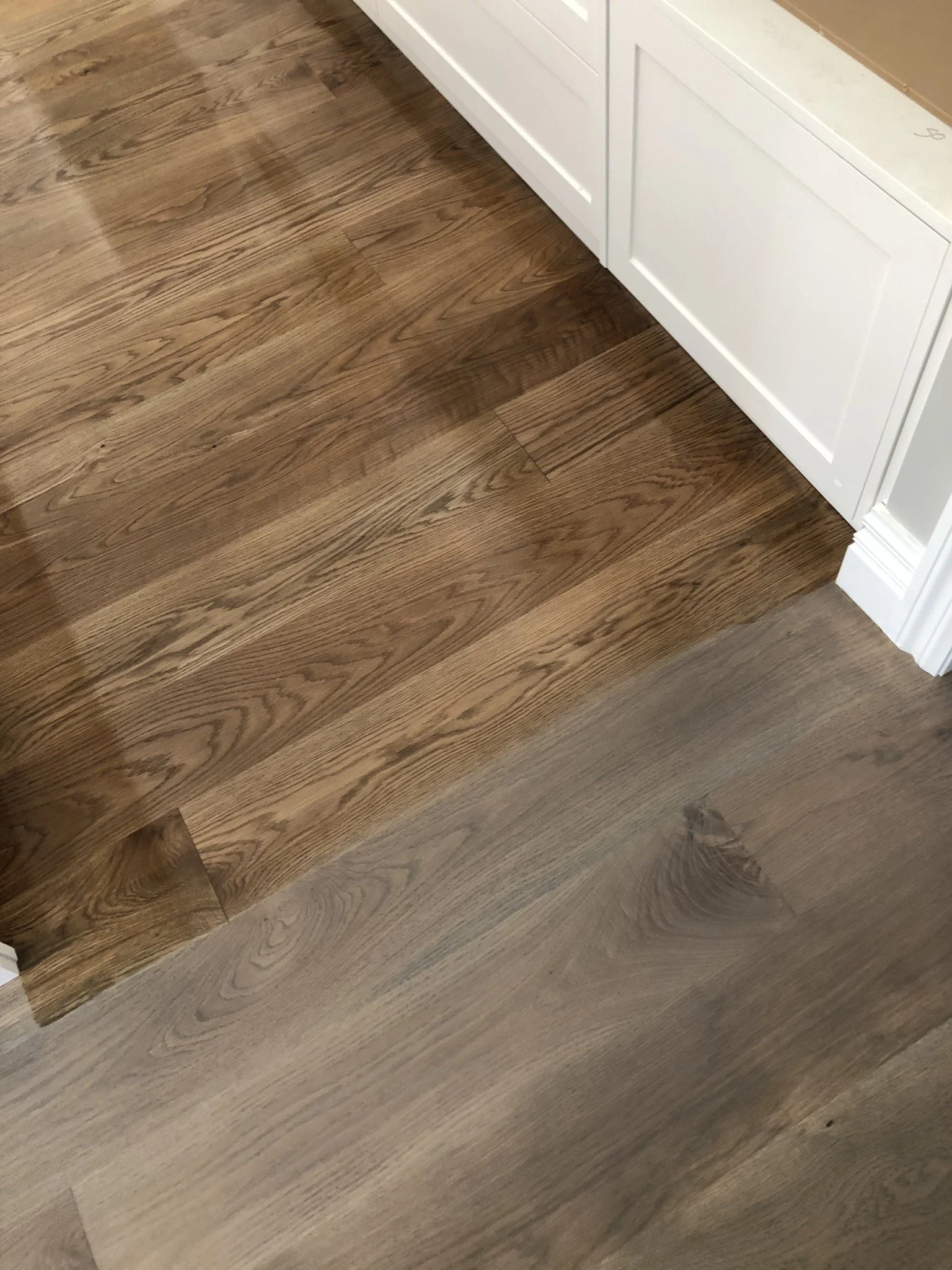 Close-up of two different hardwood floorings, one darker and one lighter, meeting at a corner near white cabinetry.