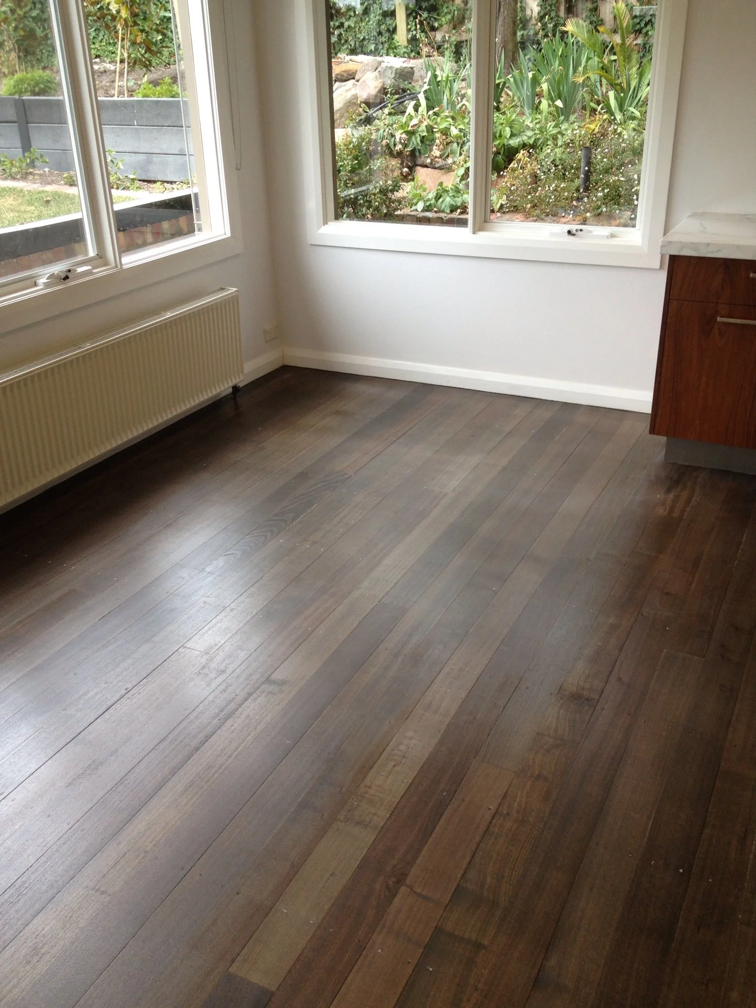 Empty room with large windows, wooden floor, white walls, and a radiator, overlooking a garden with plants and trees.