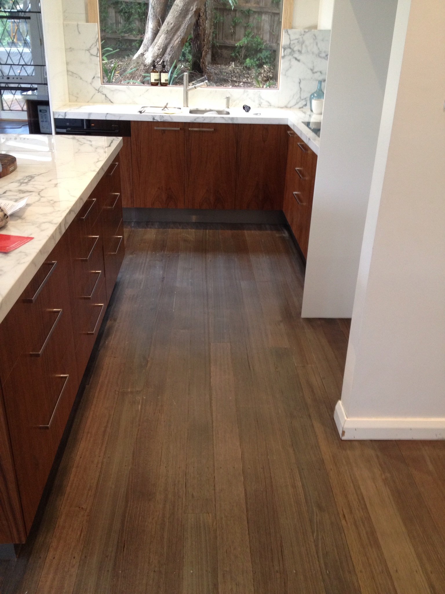 A kitchen with wooden cabinets, a marble countertop, and a large window showing a tree outside.
