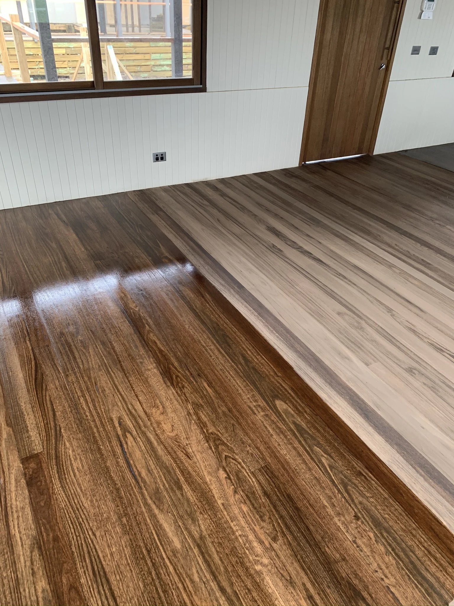 Interior of a room with newly installed hardwood flooring, showing a transition between darker and lighter wood planks, a window, and a wooden door.