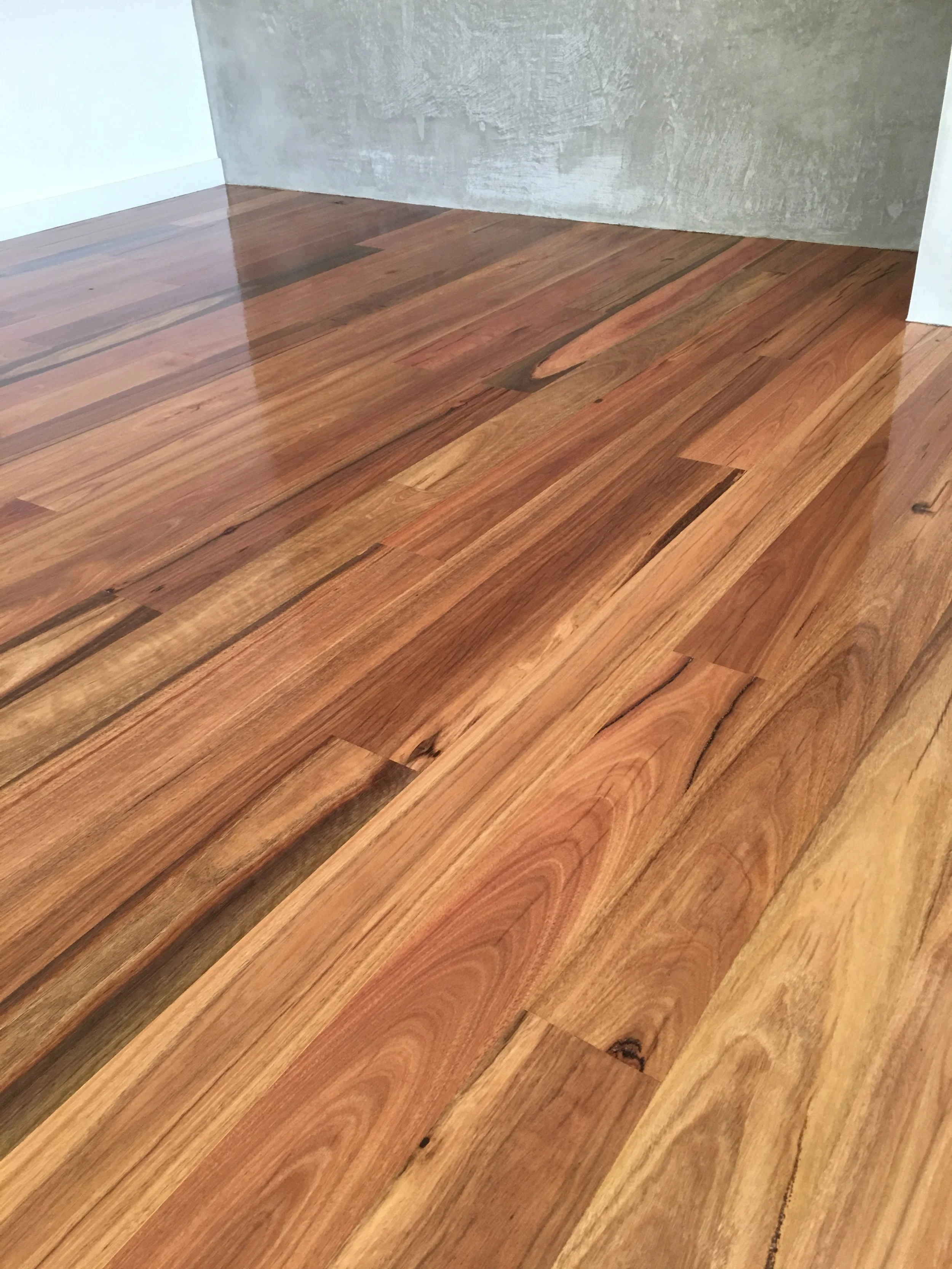 Close-up of a polished hardwood floor with natural wood grain and varying shades of brown, adjacent to a concrete wall and white baseboard.