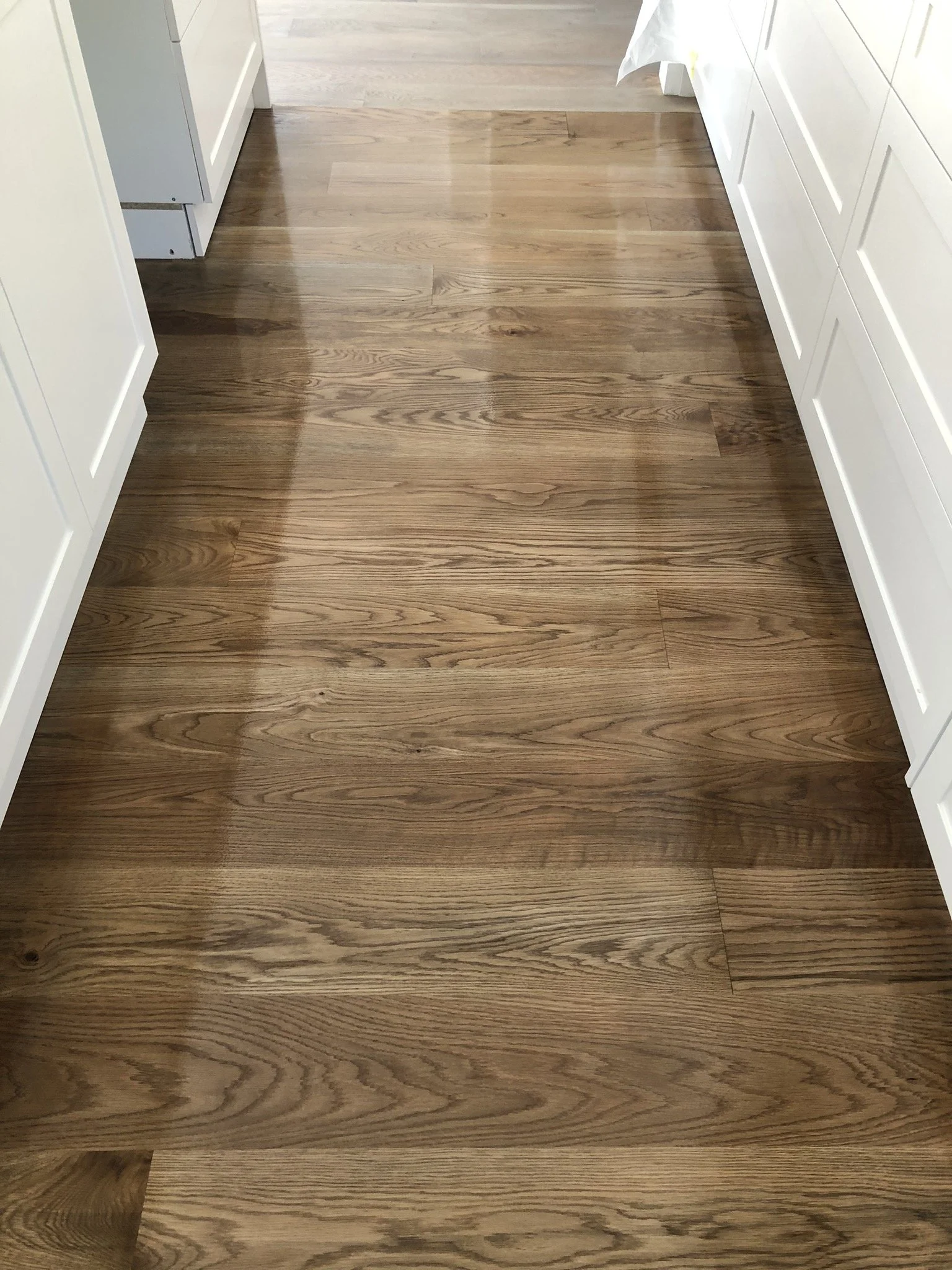 Finished hardwood floor installation in a kitchen or hallway with white cabinets on both sides.