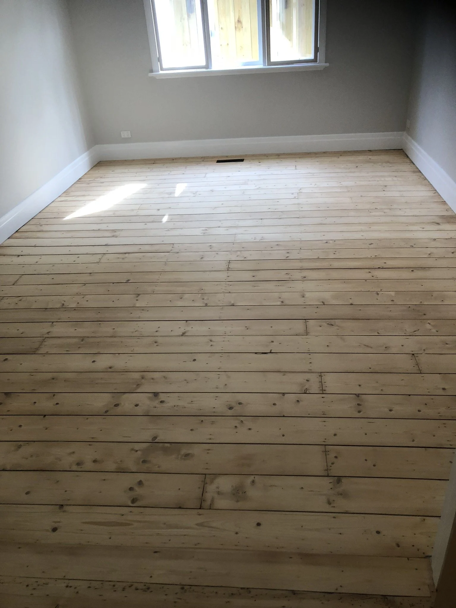 An empty room with light-colored wooden floorboards, beige walls, white baseboards, a window with sunlight streaming in, and a floor vent near the window.