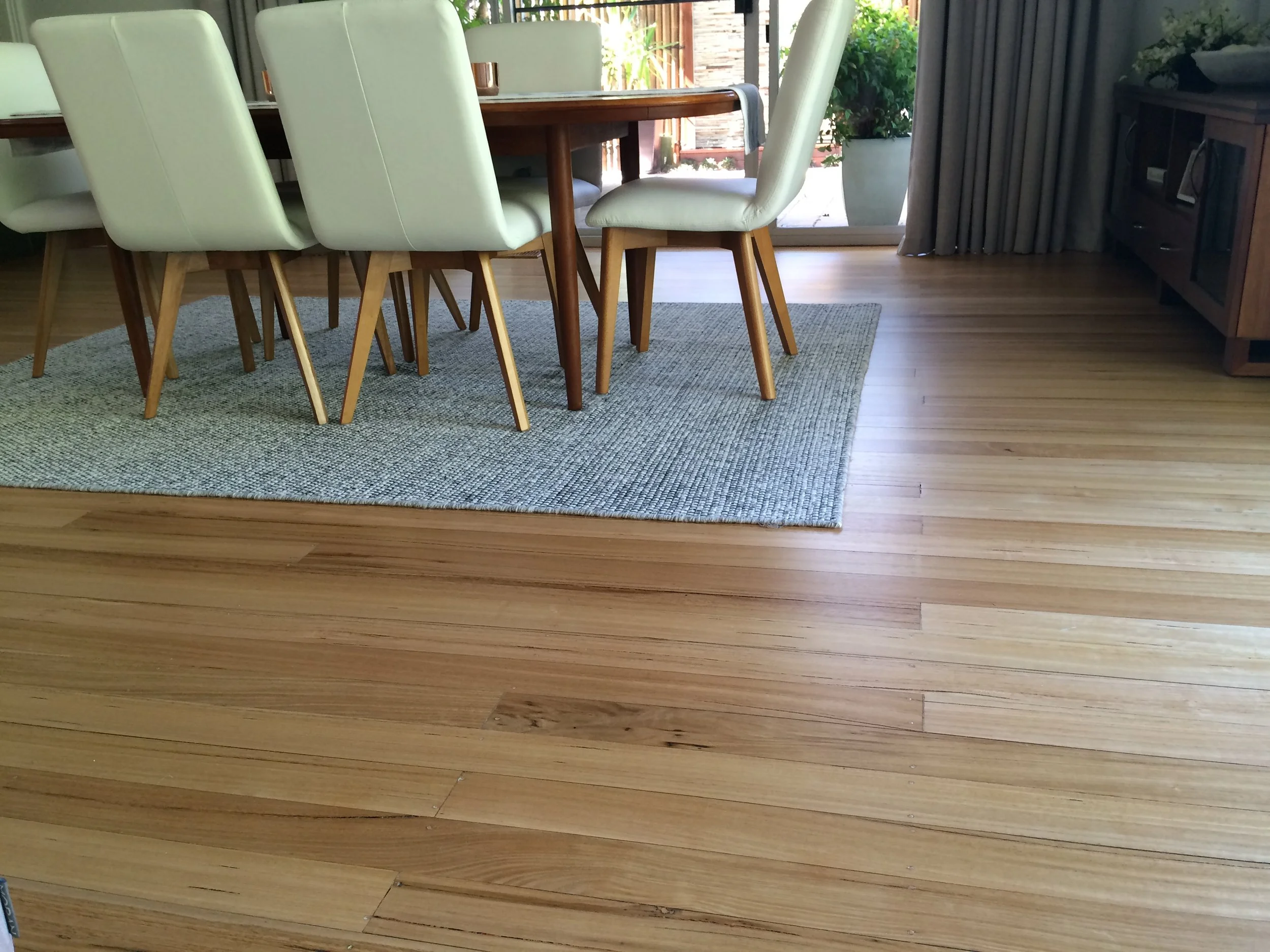 A dining room with a wooden table surrounded by eight white upholstered chairs with wooden legs. The floor is hardwood, and there is a gray rug under the table. In the background, large glass sliding doors with gray curtains reveal an outdoor patio w