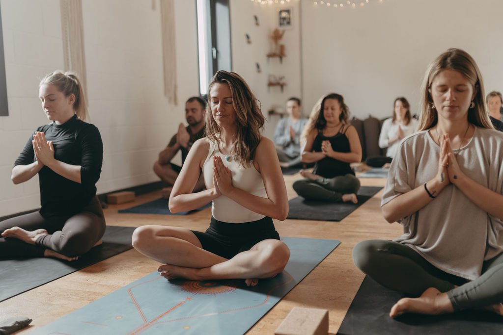 Group of women and one man practicing yoga or meditation in a cozy room with wooden floors, sitting cross-legged on mats with eyes closed and hands in prayer position.