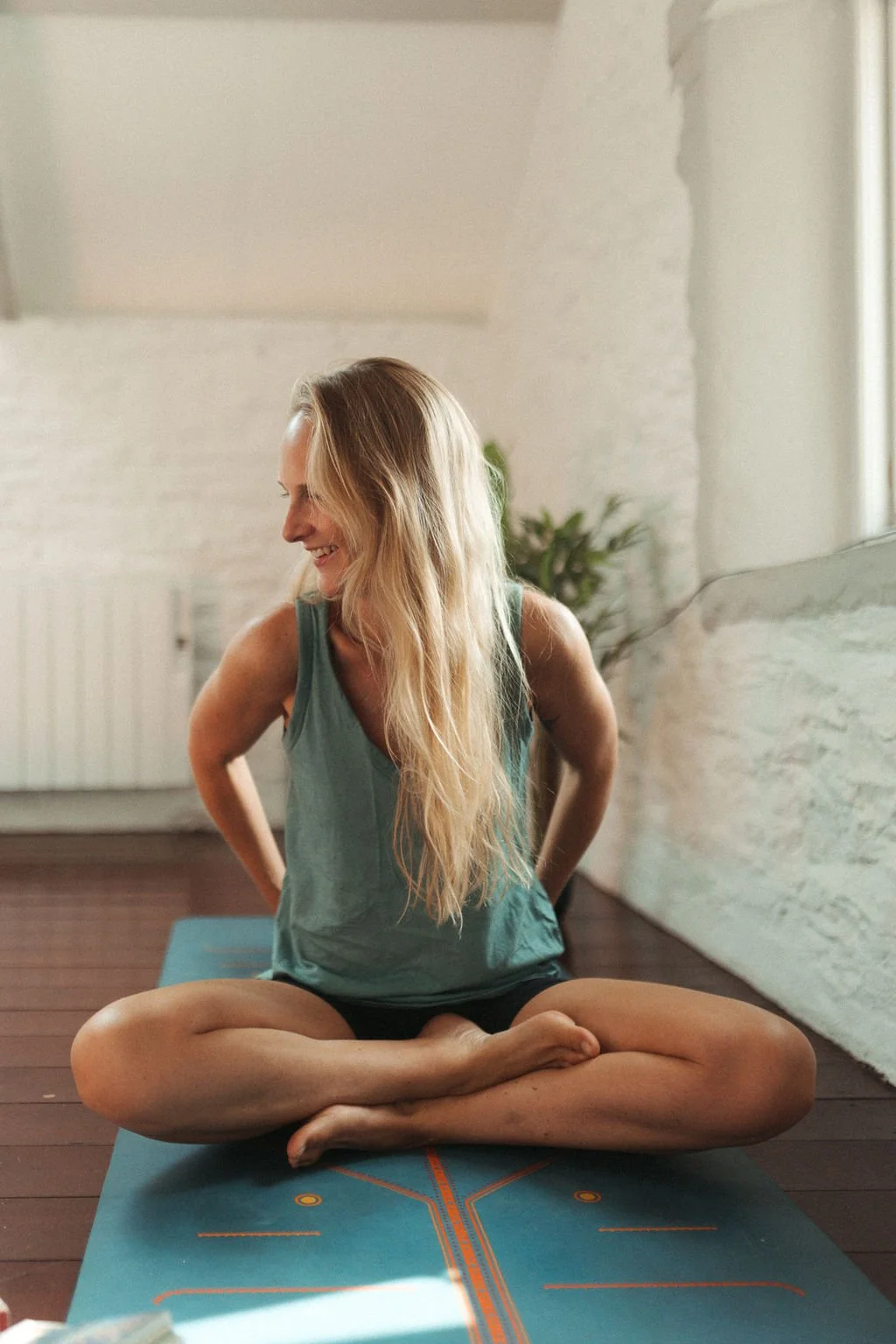 A woman with long blonde hair sitting cross-legged on a yoga mat in a bright room, smiling and looking down.