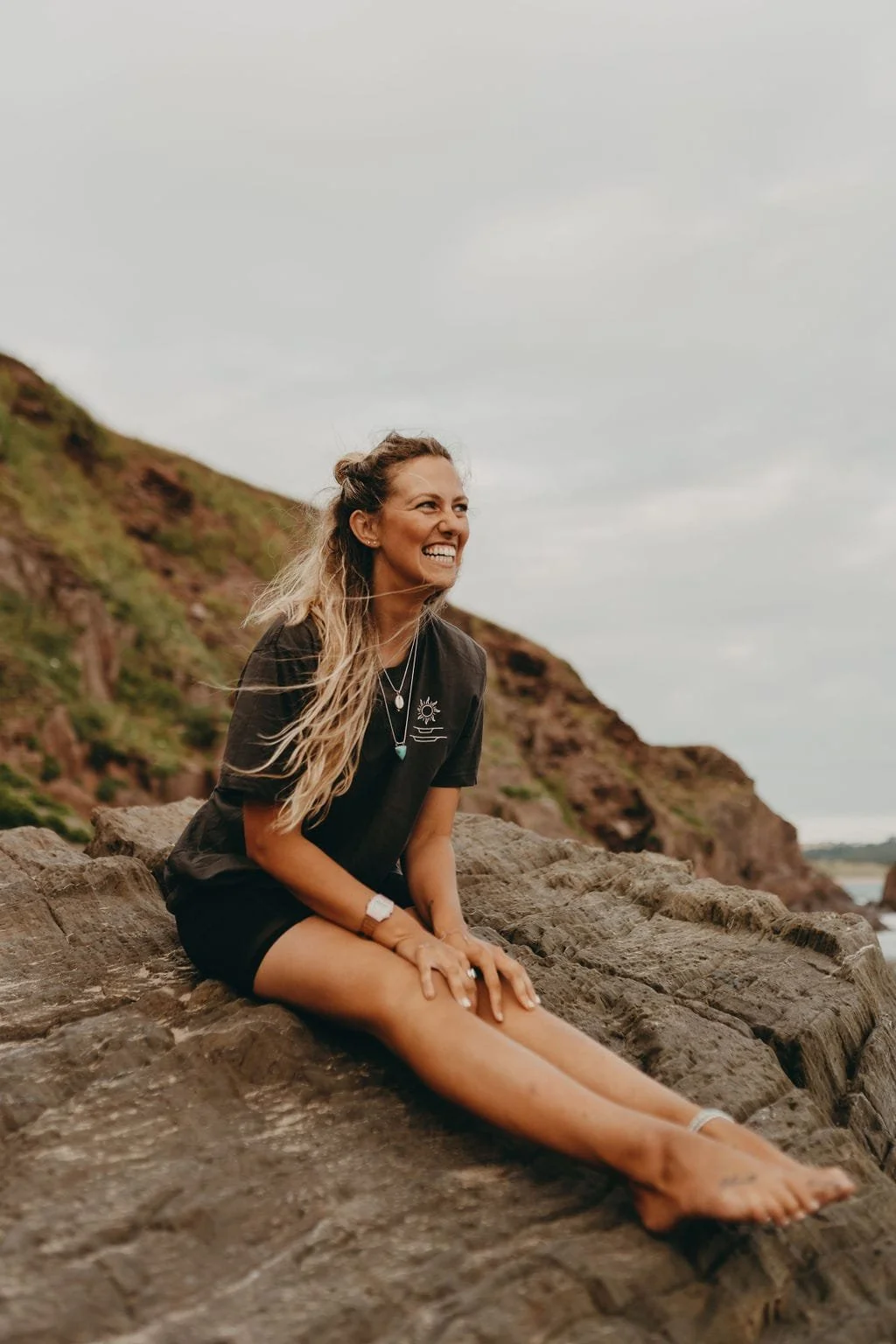A woman with long, wavy hair sitting on a rock by the beach, smiling and enjoying her time outdoors near a hillside under a cloudy sky.