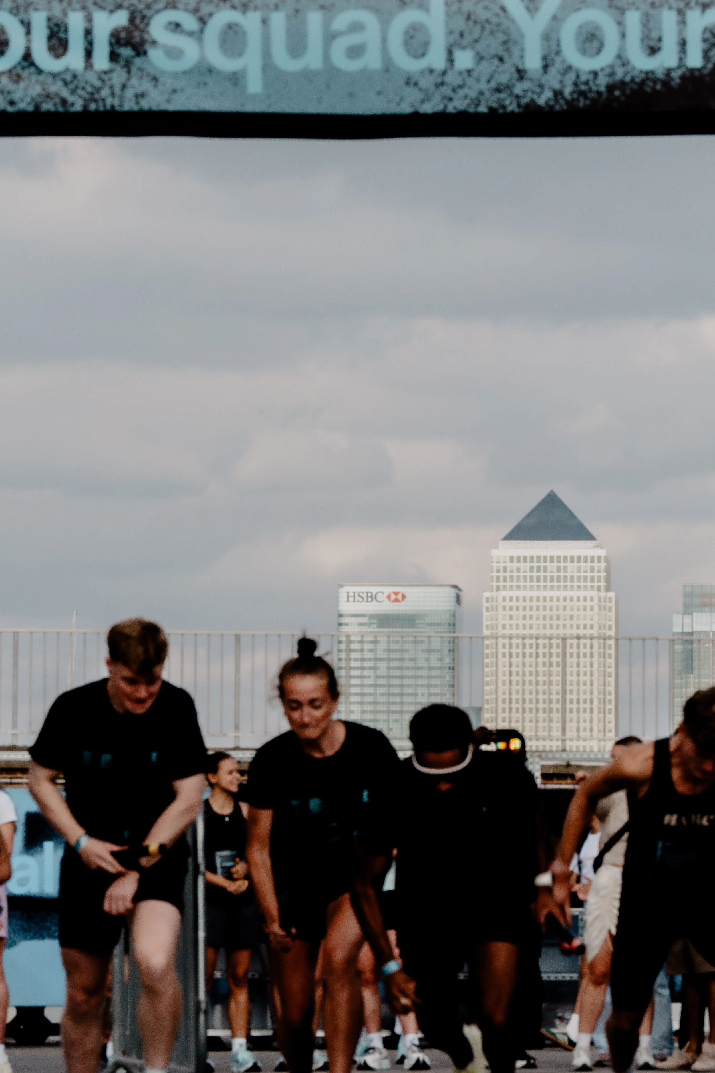 Group of people wearing black shirts engaging in a workout session or race on a rooftop, with a city skyline in the background, including tall buildings and cloudy sky.