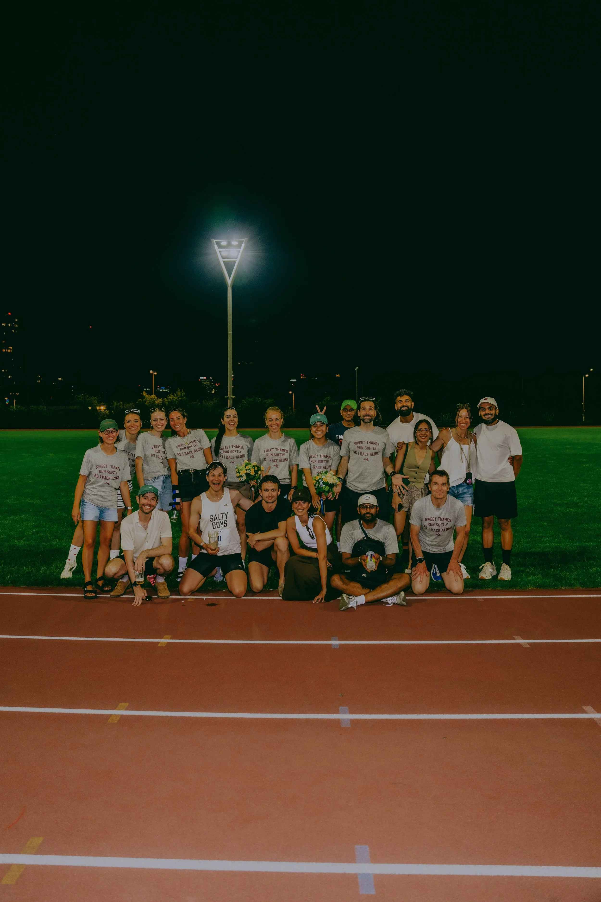 A group of people gathered on a track at night, with a grassy field and city lights in the background, posing for a photo.