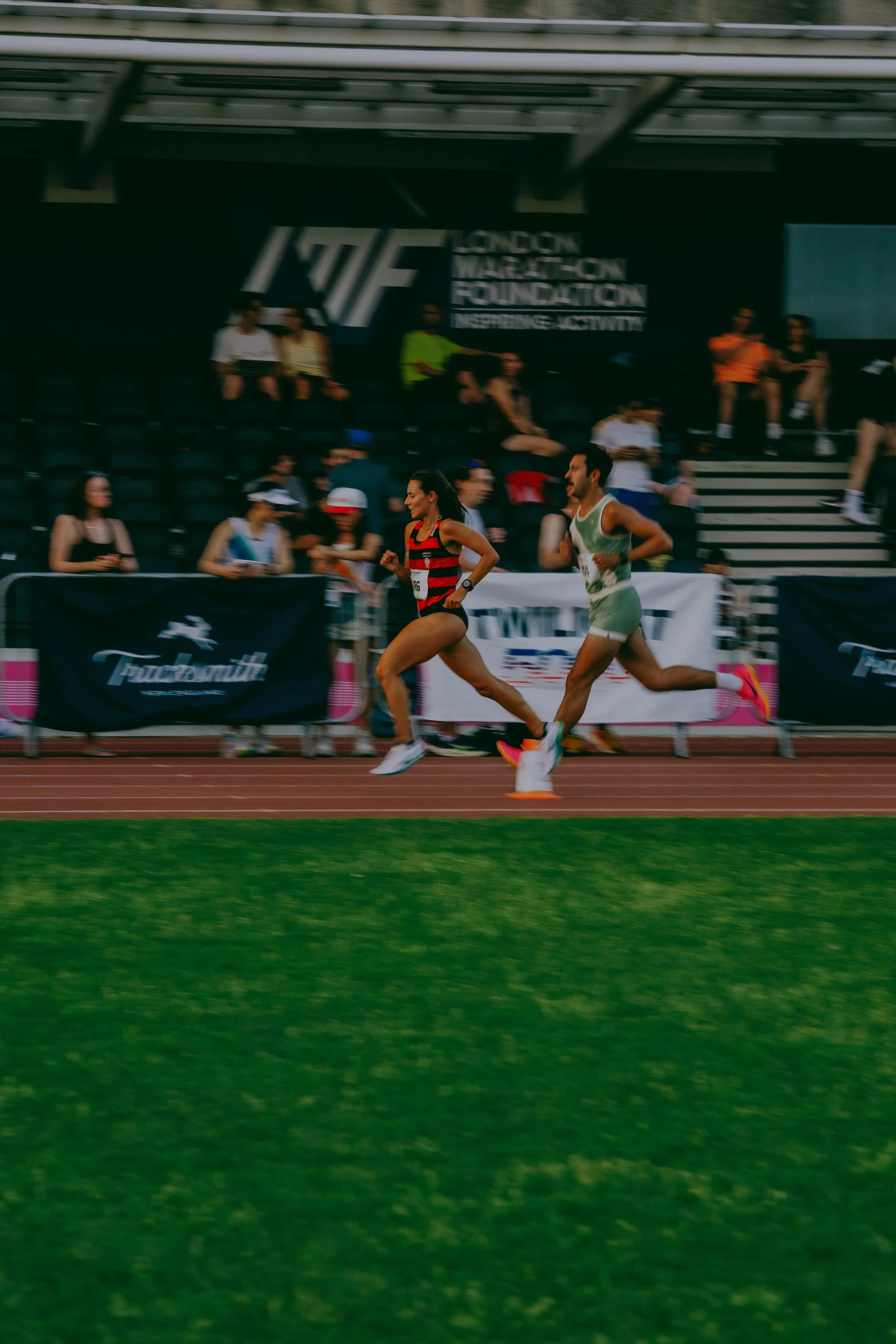 Two runners, a woman in a red and black striped outfit and a man in a green and white outfit, competing in a race on a red and green track, with spectators watching from the stands and banners in the background.