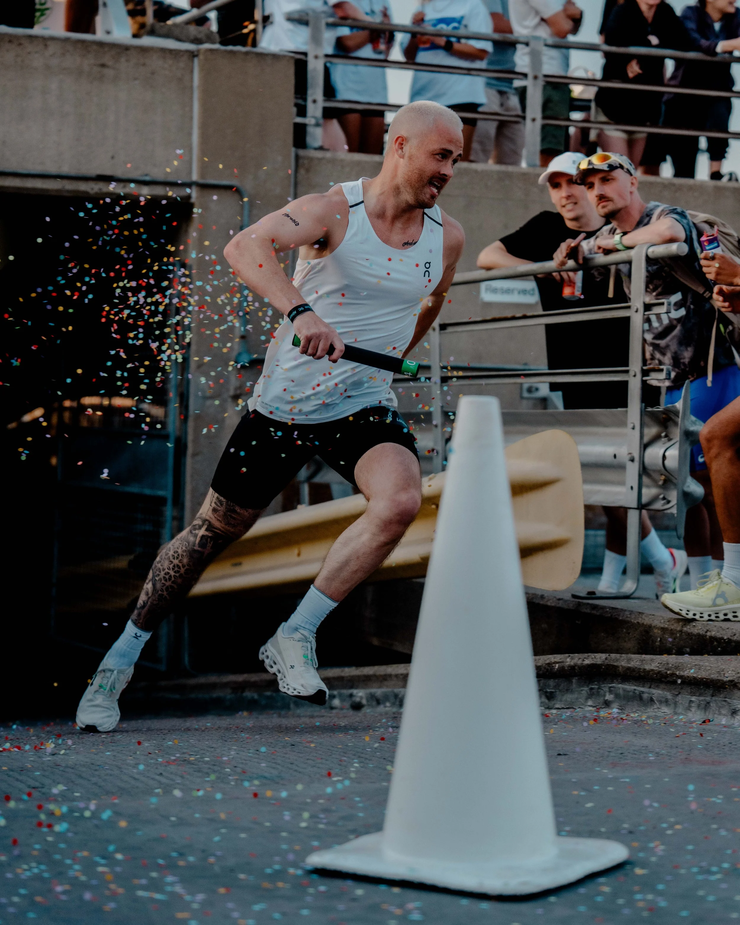 A male runner crossing a finish line, surrounded by colorful confetti, with spectators and supporters watching from behind a metal railing.