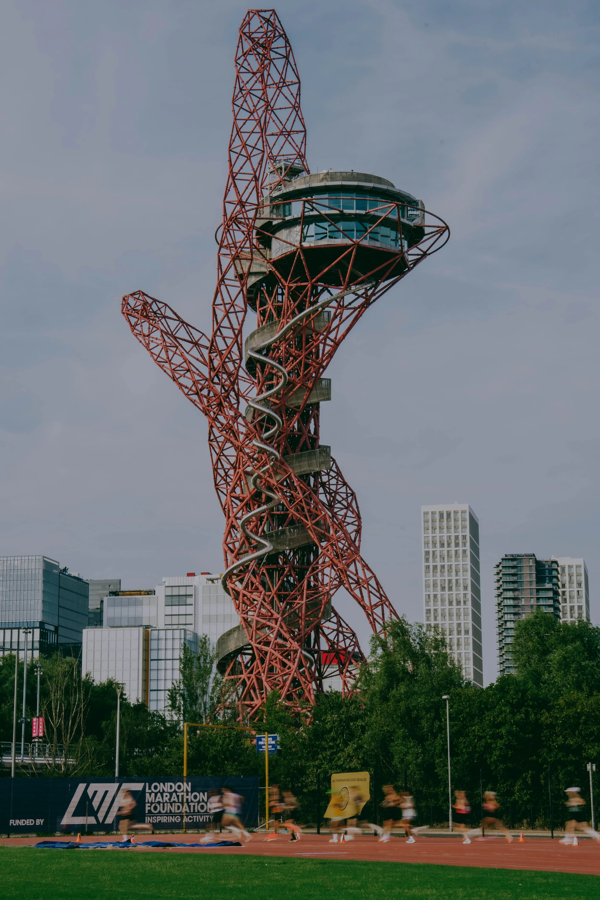 The image features the ArcelorMittal Orbit, a red metal sculpture and observation tower with a spiral slide, set in an urban park. In the foreground, runners are participating in a race on a track, with a banner reading 'London Marathon Foundation' a
