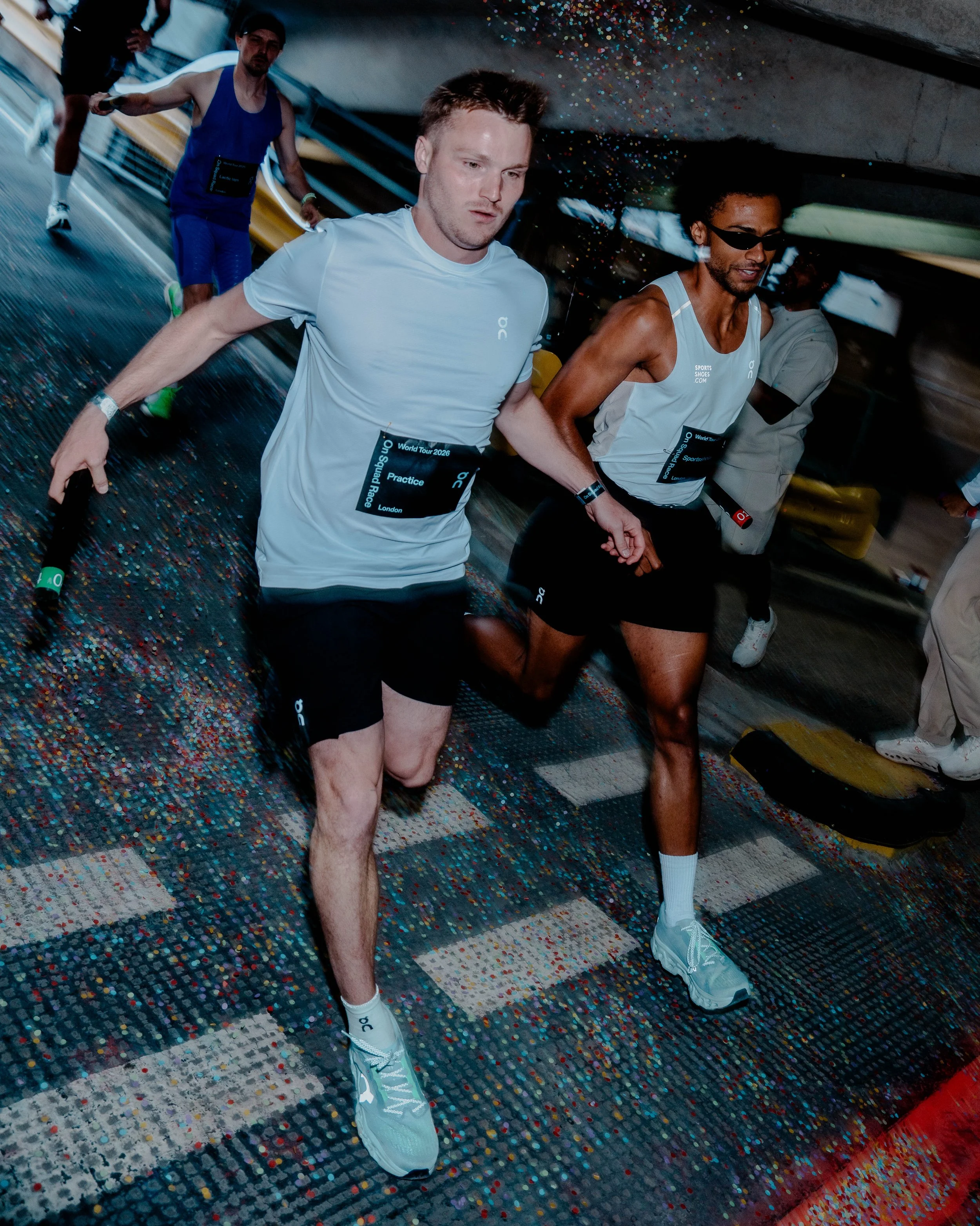 Three male runners in athletic gear participating in a race indoors, with a colorful, speckled floor and a concrete wall in the background.