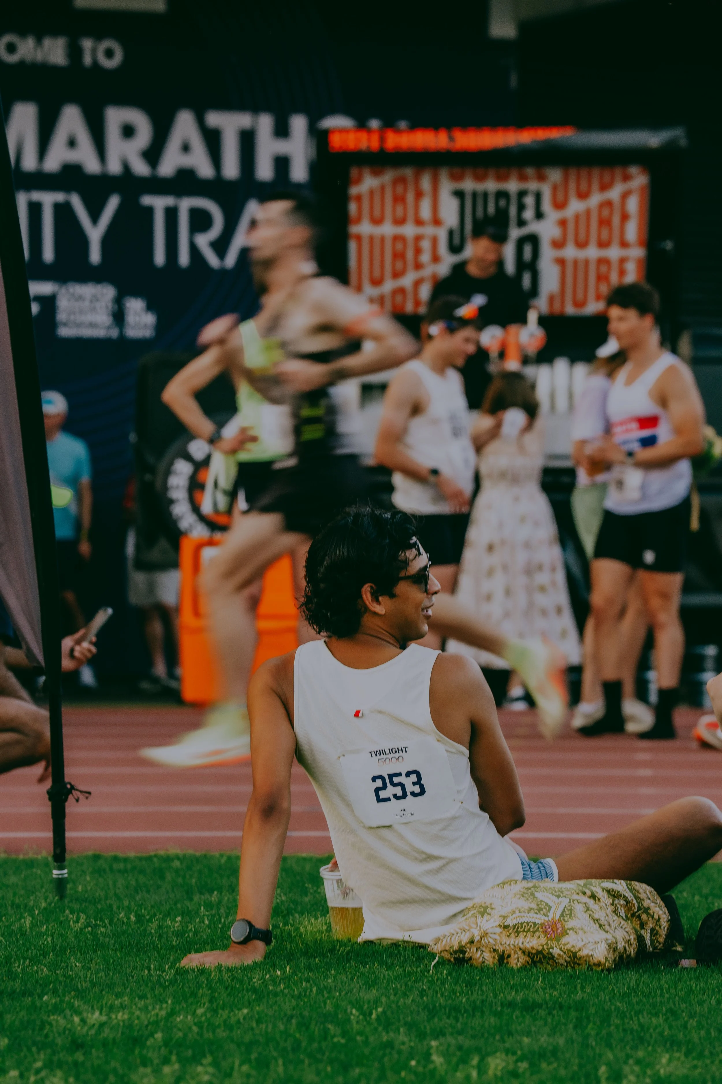 A person with a race bib sitting on the grass at a track and field event, with other athletes and event signage in the background.