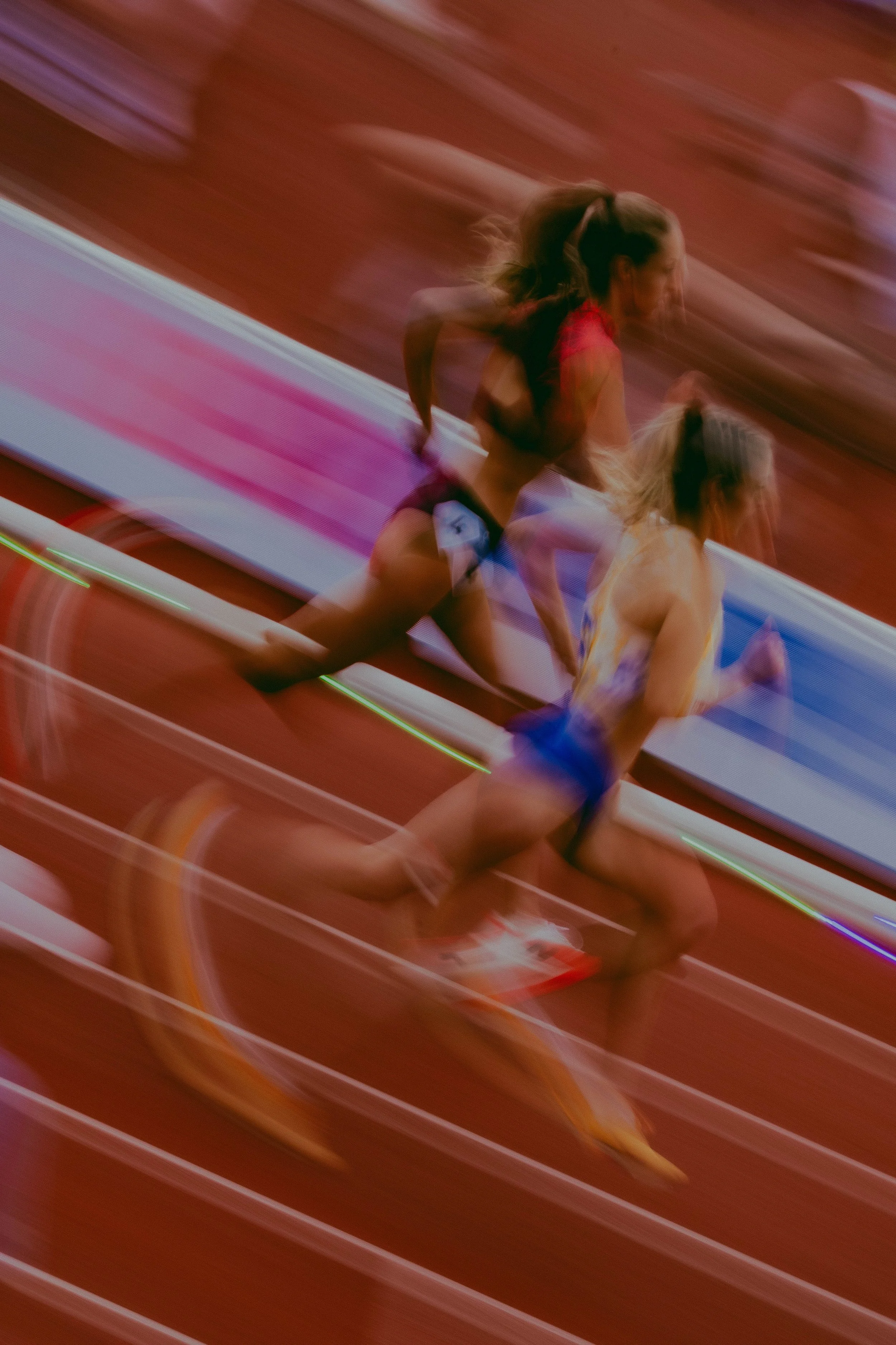 Two female runners on a red indoor track, captured with motion blur.