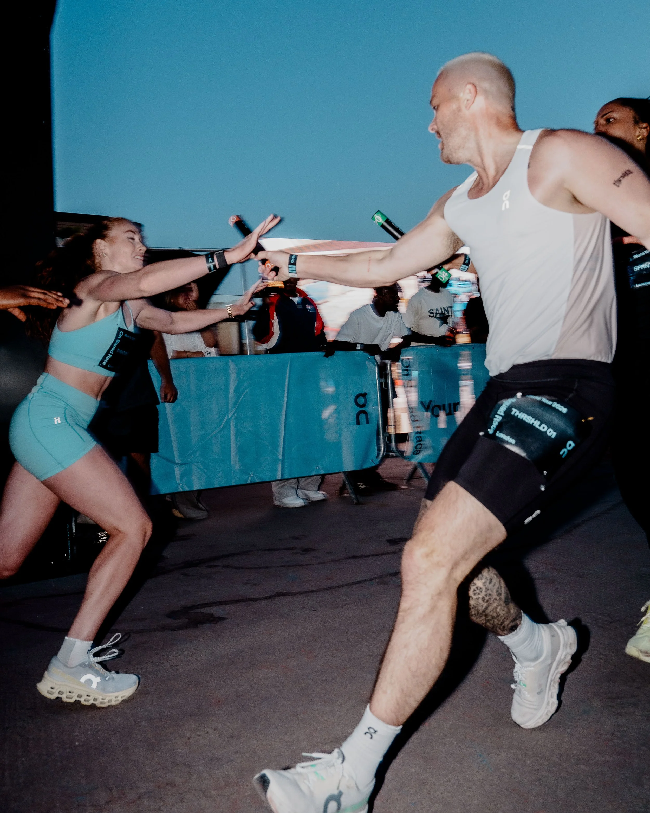 A female athlete in a blue sports outfit reaching out to accept a high five from a male athlete in a white tank top, with other people and event elements visible in the background, during a running event.