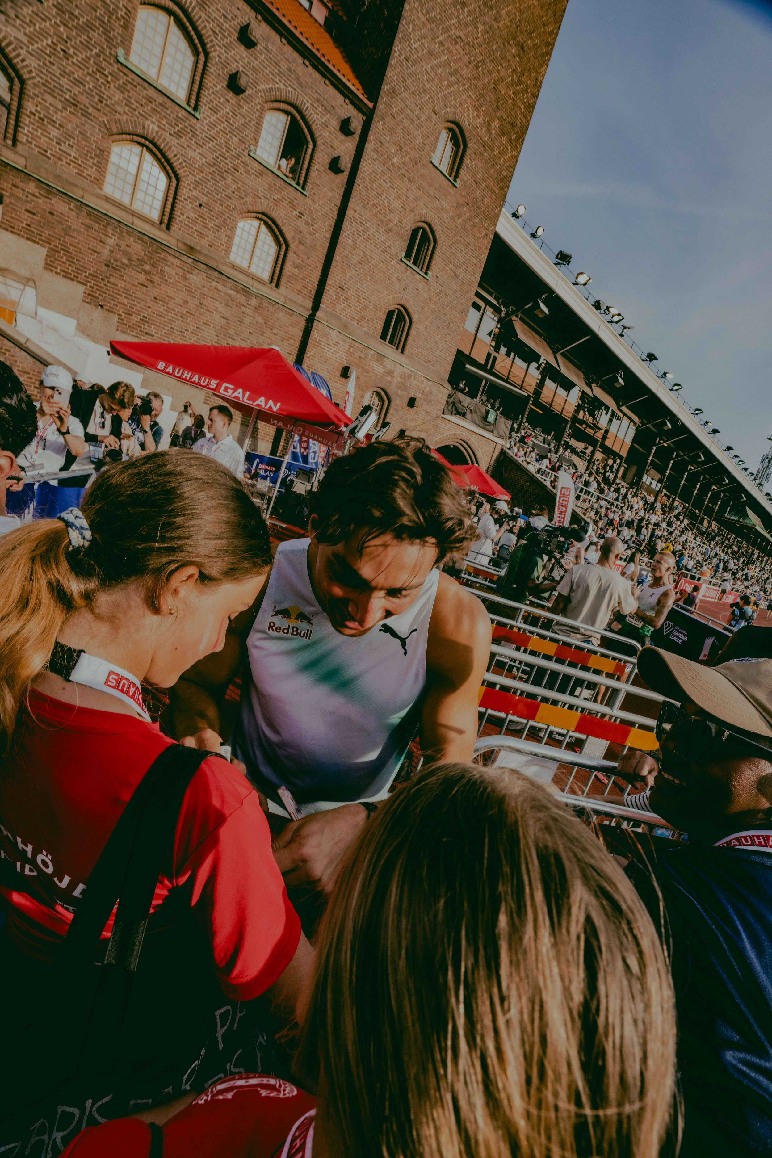 Athletes and spectators at a sports event in front of a historic brick building, with some people taking photos and engaging with athletes under a red canopy marked "Bauhaus Galan."