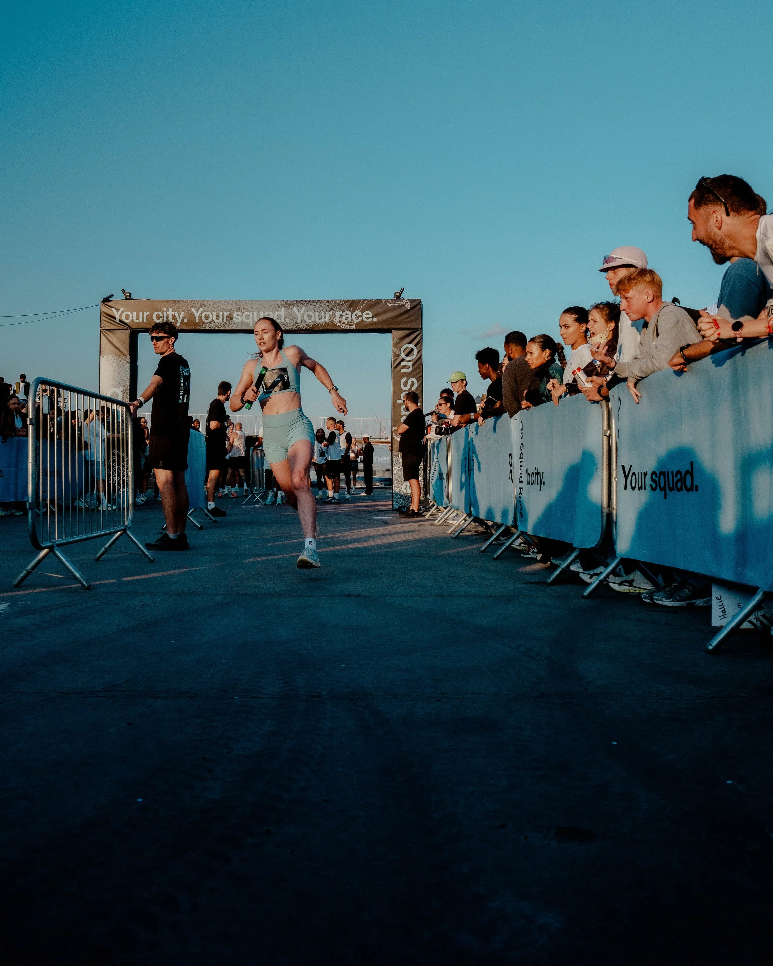 A woman crossing the finish line at a race, surrounded by spectators and event banners on a clear day with a bright blue sky.