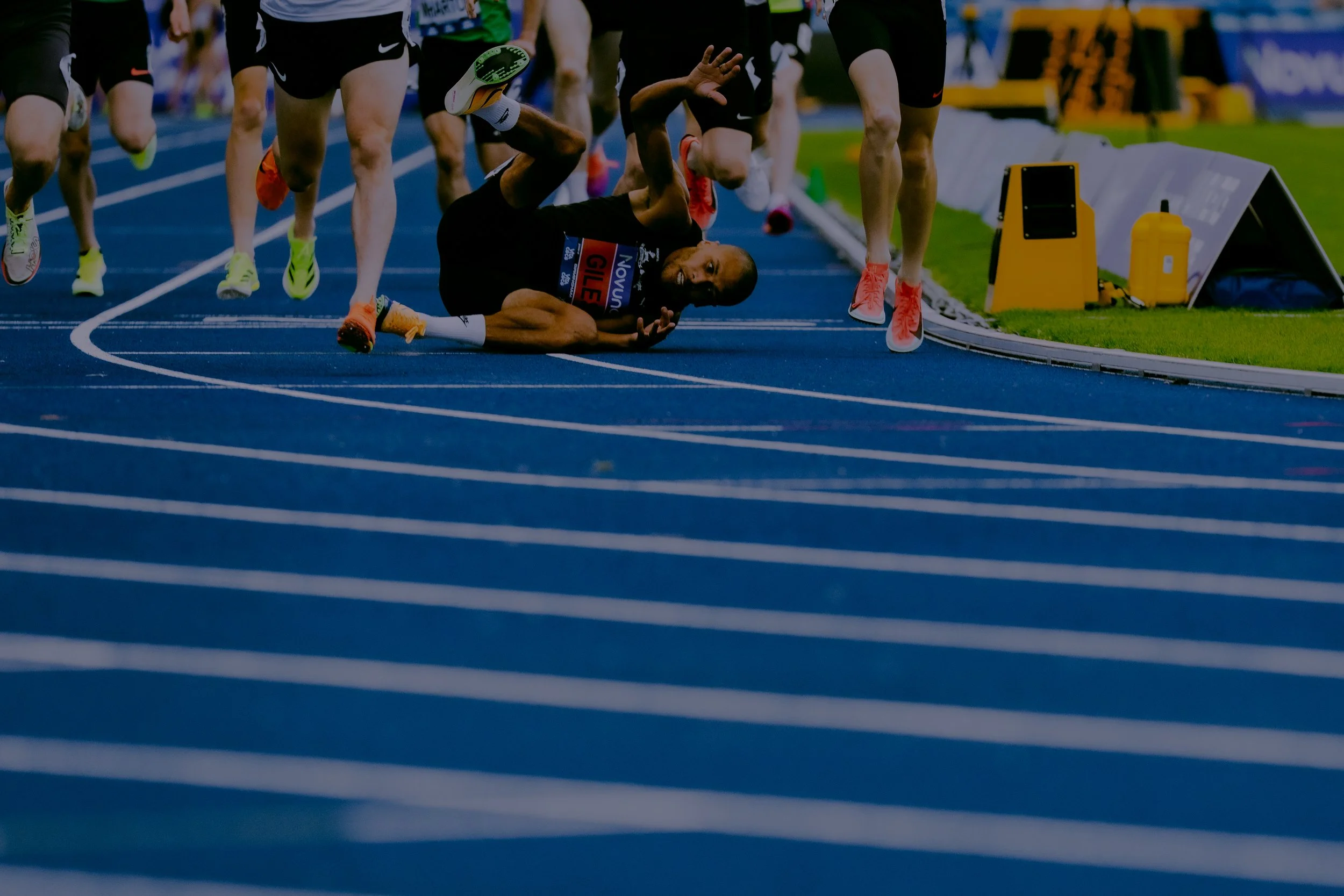 A runner has fallen on a blue track during a race, with several other runners around him. The fallen runner is on his side, smiling, and holding his arm up while lying near the track's edge. The other runners are standing or running past, and there a