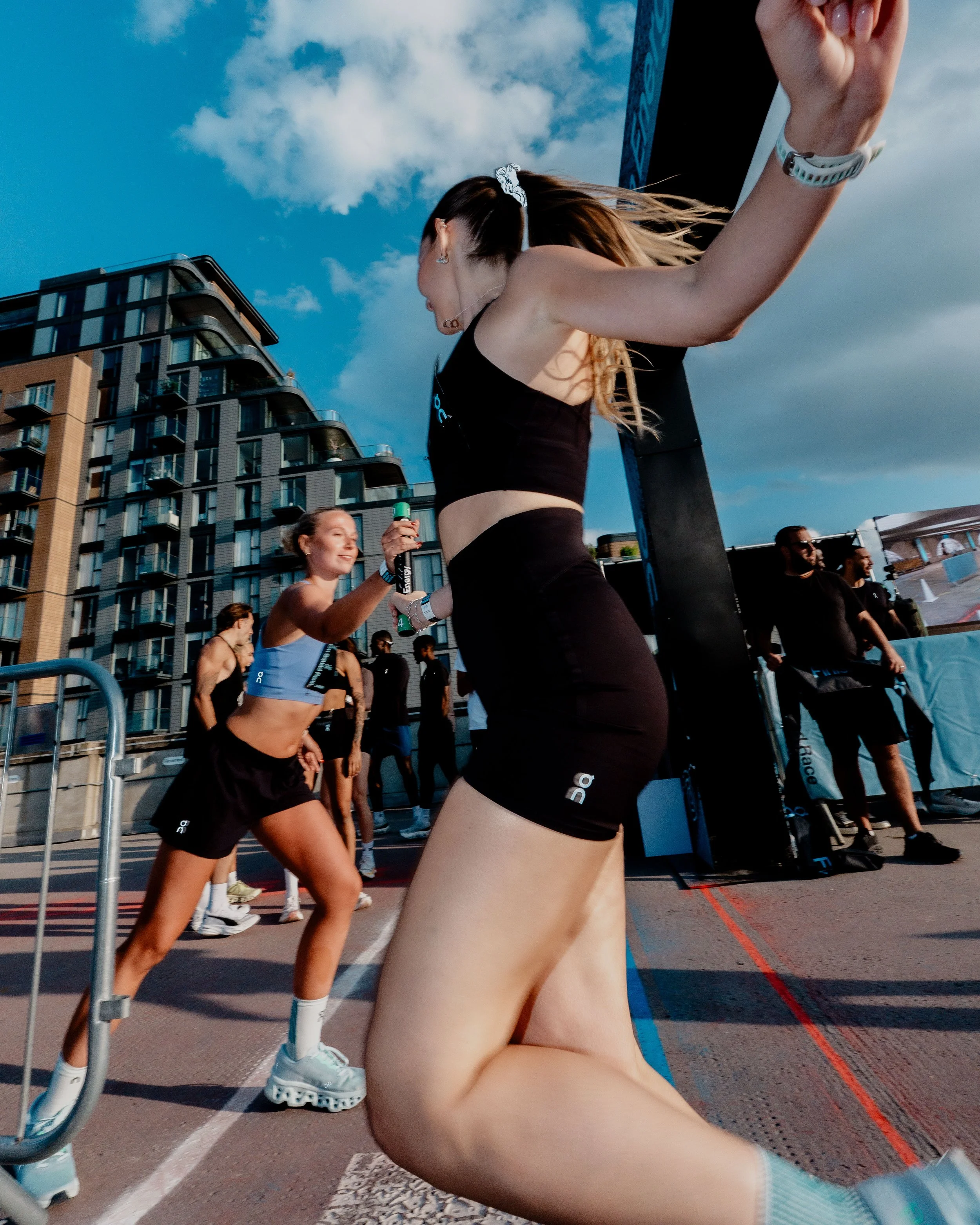A group of people running in an outdoor race, with a woman in black athletic wear kneeling on the ground in the foreground, and other runners and a building with balconies in the background on a partly cloudy day.