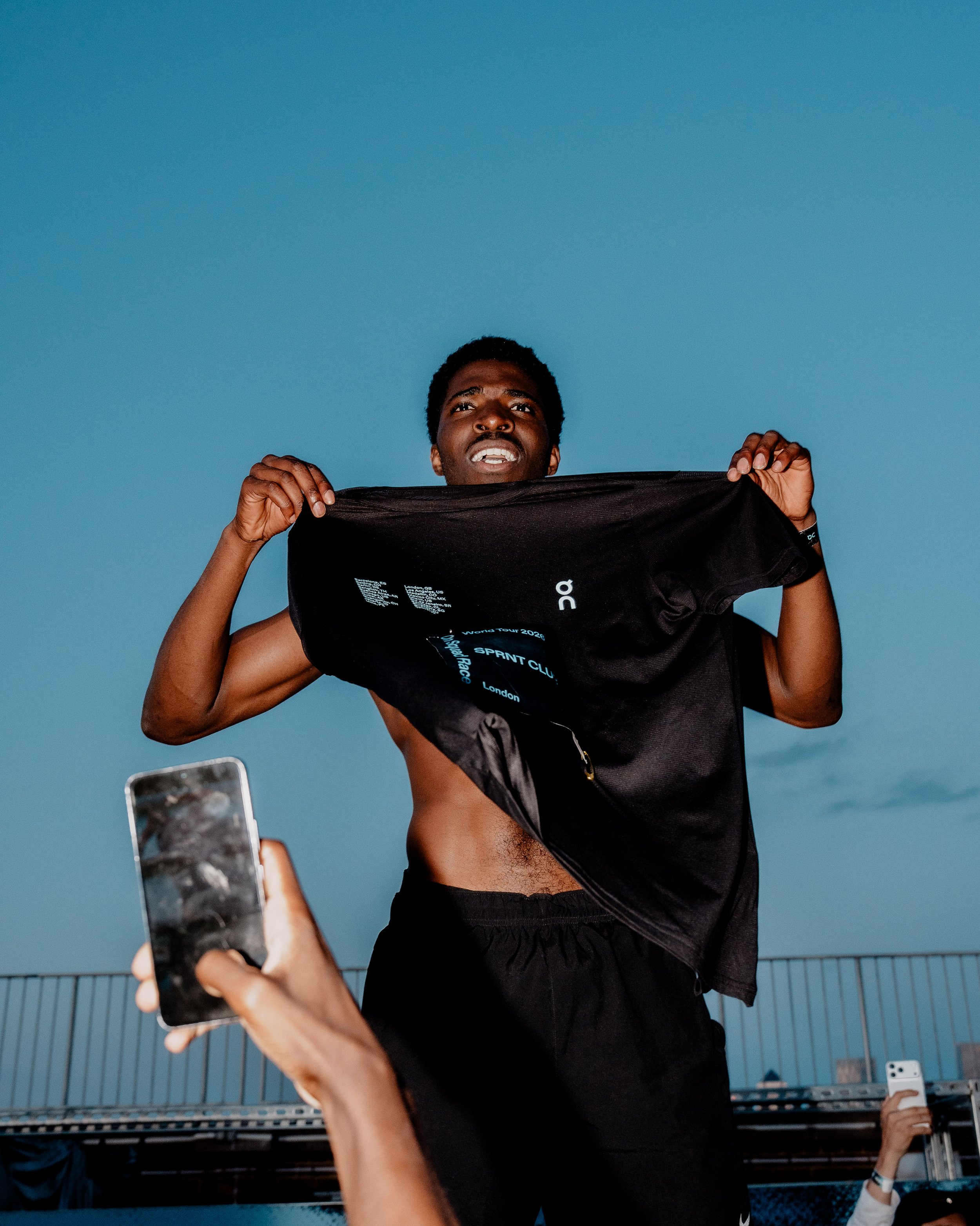 A man holding a black t-shirt, with another person in the background taking a photo with a smartphone, on a rooftop with a blue sky.