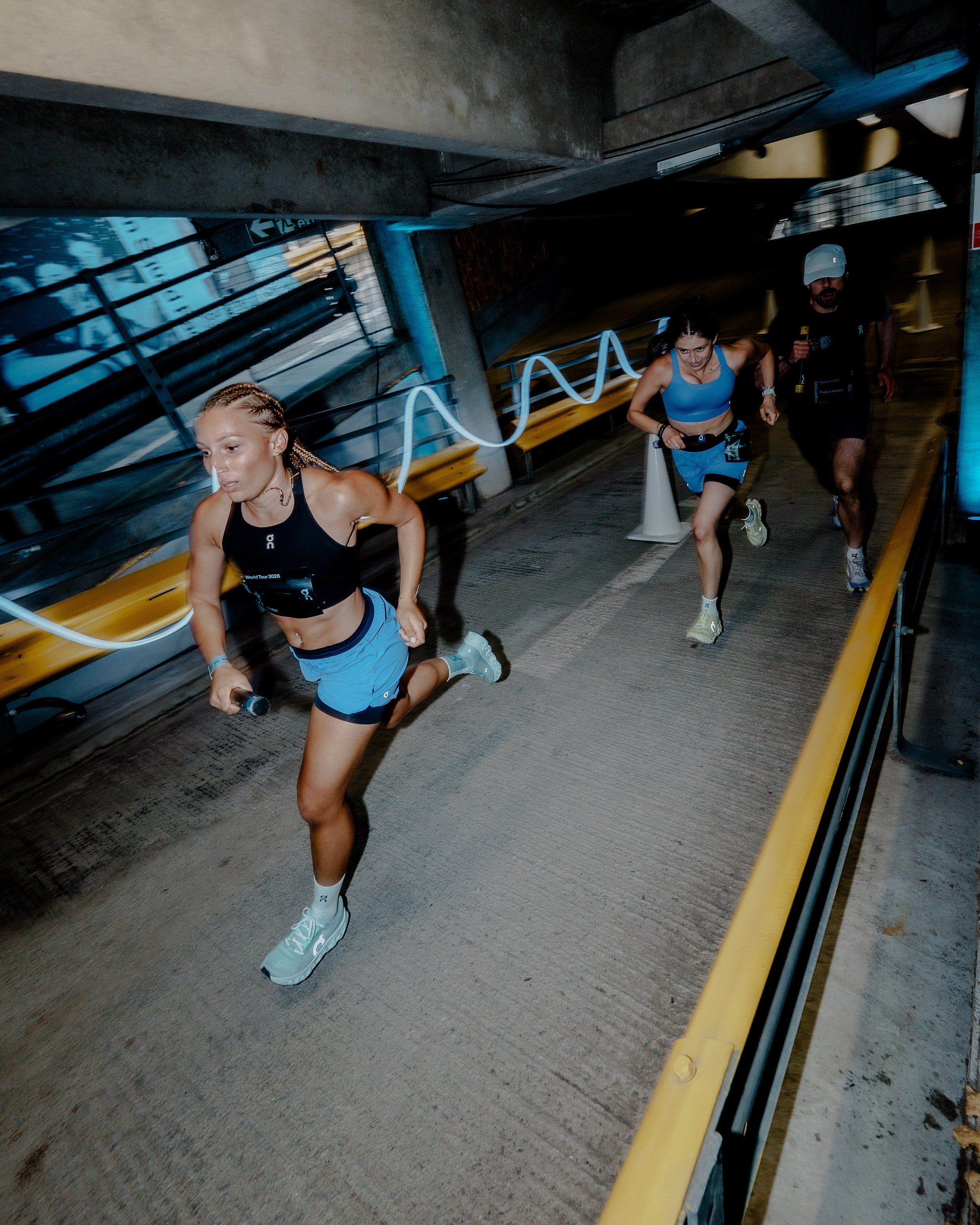 Three runners participate in a race, running in an underground tunnel with concrete walls, metal railings, and a yellow safety barrier, with the woman in the front holding a phone.