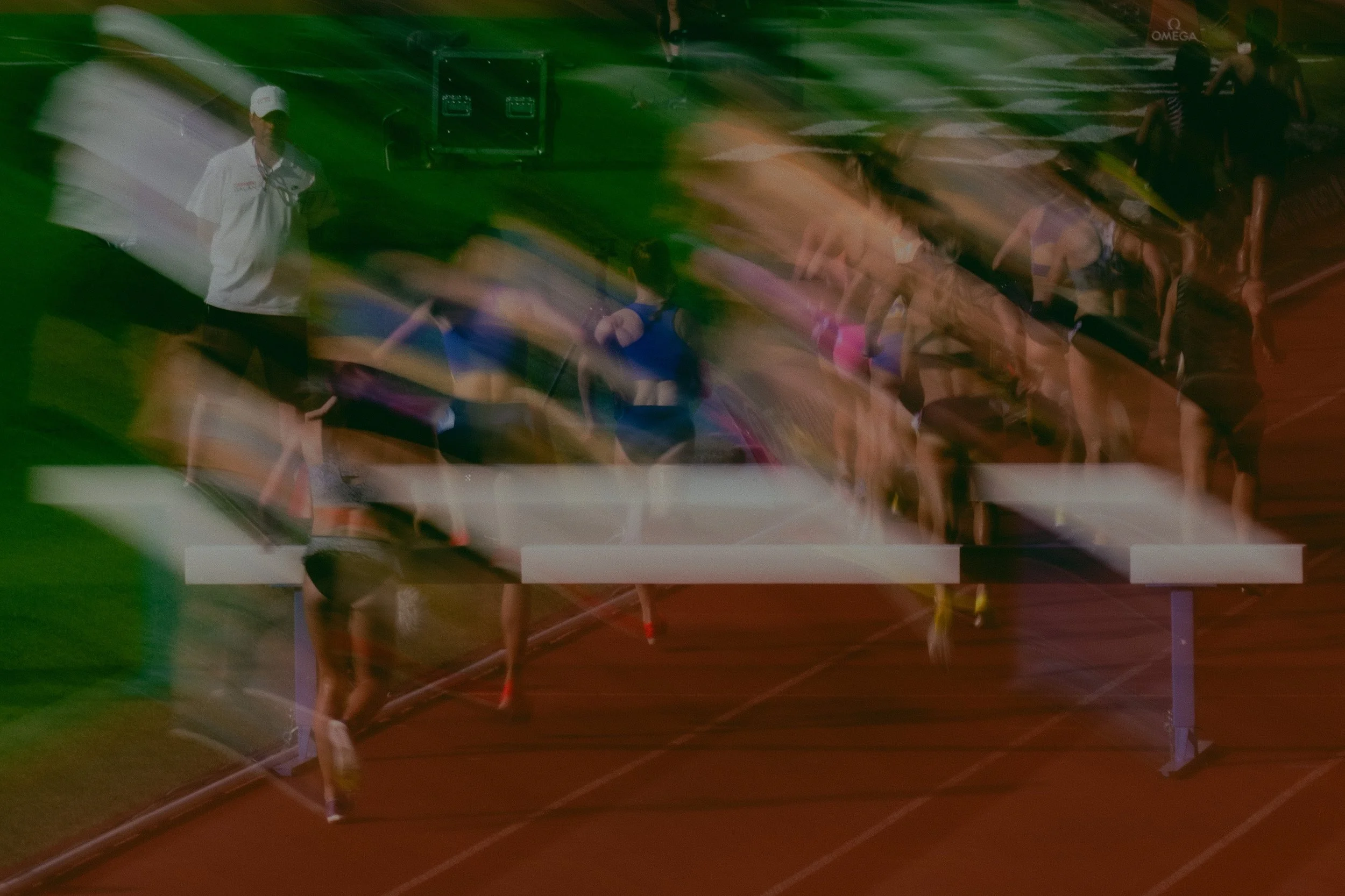 Man in white shirt and cap standing near a track field with multiple people exercising or stretching on the track. The image appears blurry and distorted with colored streaks.