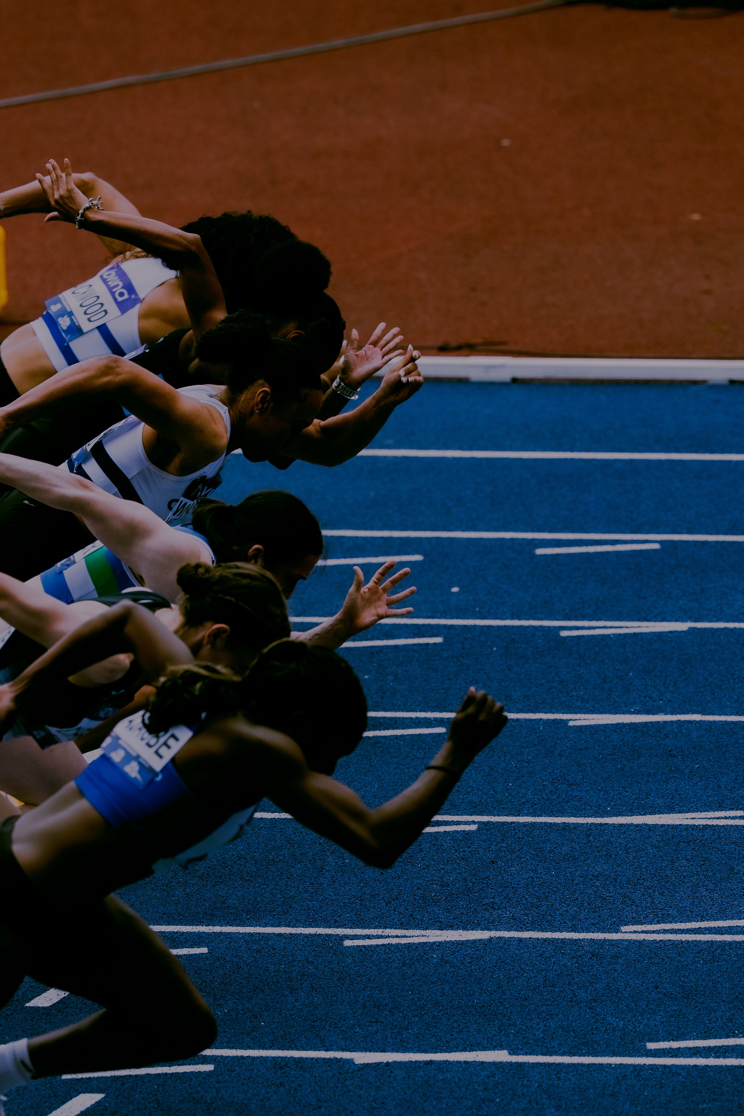 Close-up of female athletes at the start of a race on a blue track, with some athletes in athletic gear and numbered bibs, preparing to run.