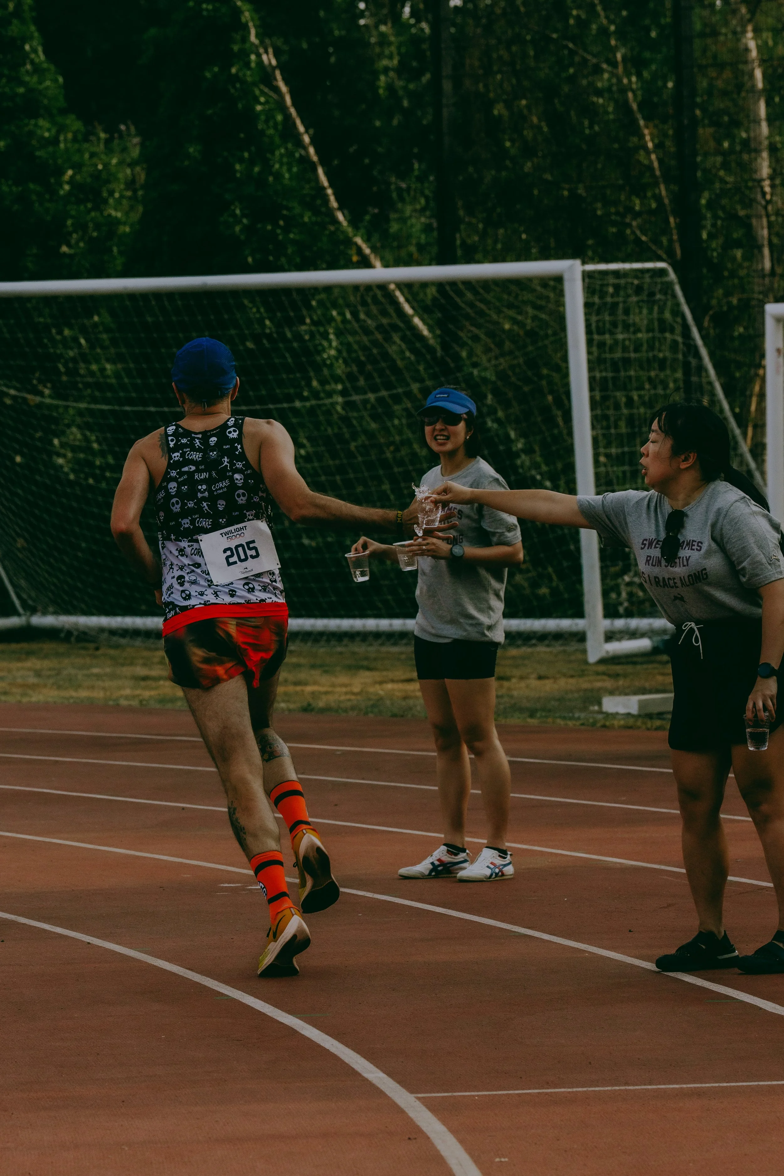 A male runner in a black skull print tank top and red shorts approaching a water station at a race, with two young women handing out water on a track surrounded by greenery.