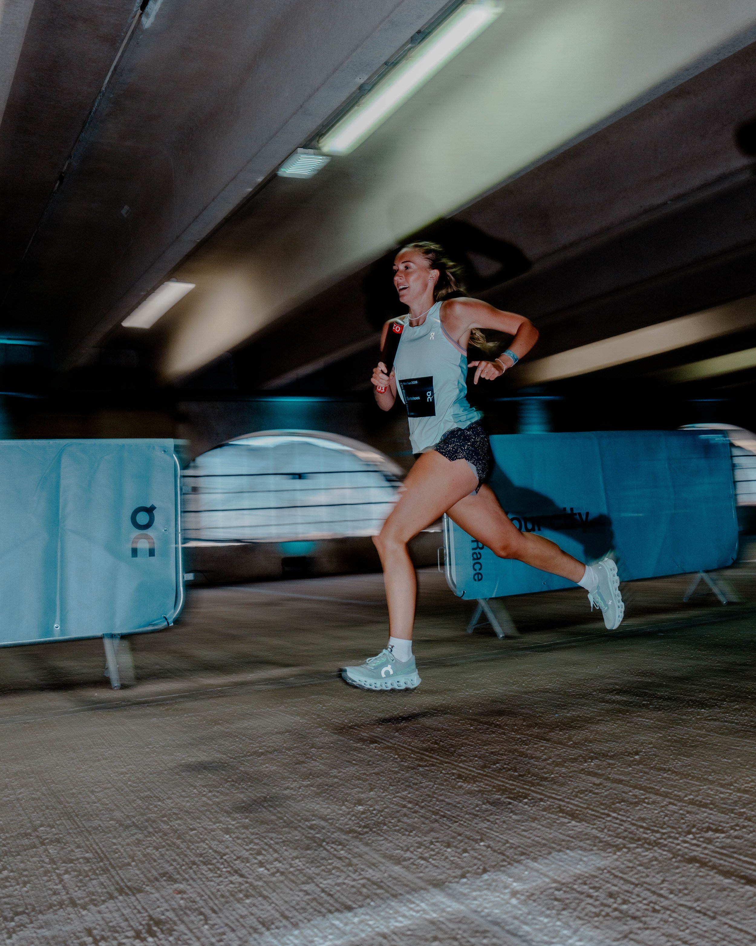 A young woman running in an indoor venue, wearing athletic gear, with banners and benches nearby, under a concrete ceiling and city lighting reflections on the floor.