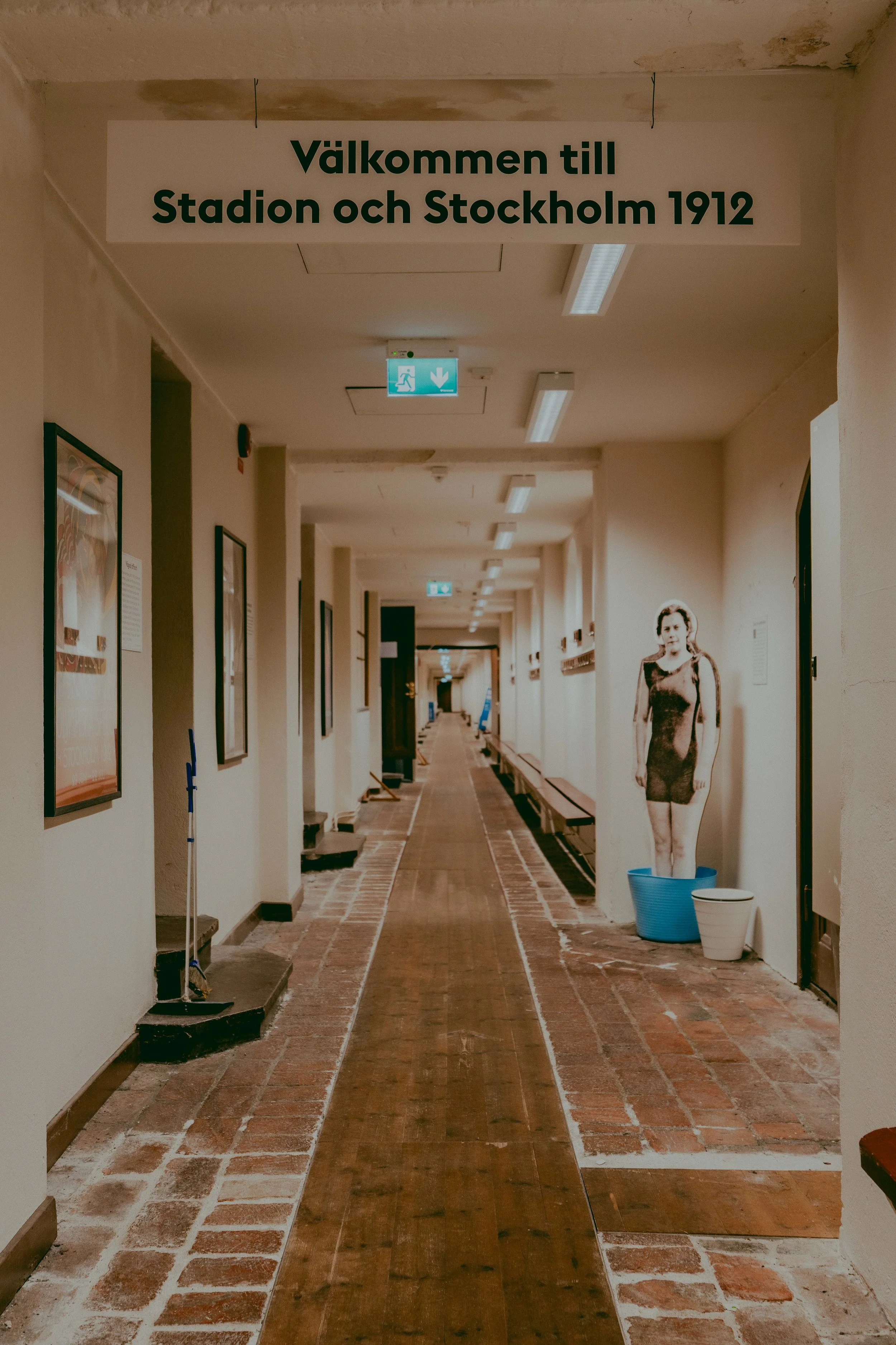 Long hallway in a building with framed posters on both sides, brick-tiled floor, and a cardboard cutout of a woman in a dress near the wall on the right, with blue and white flower pots on the floor nearby.
