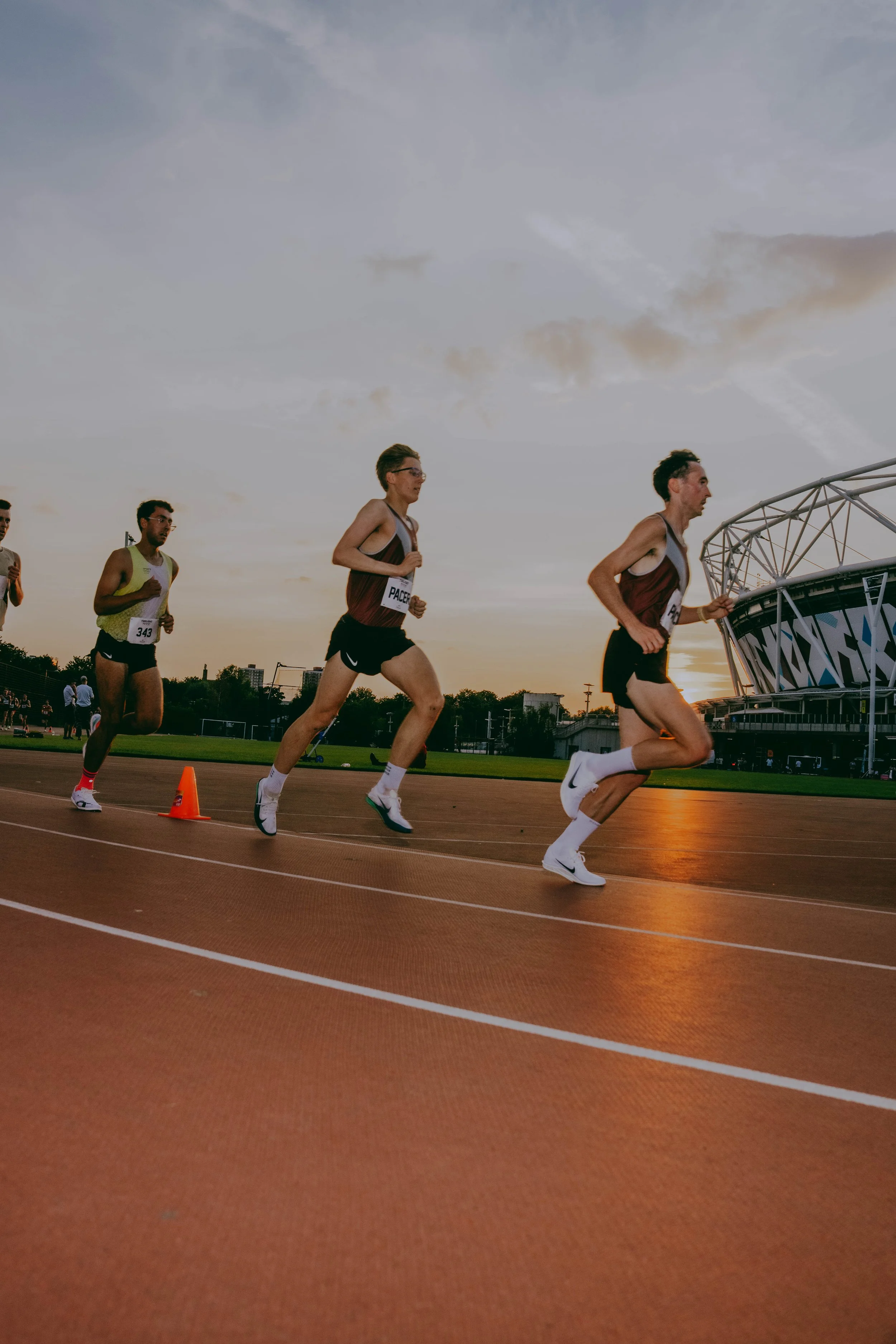 Four male runners competing in a race on a track during sunset, with stadium and buildings in the background.