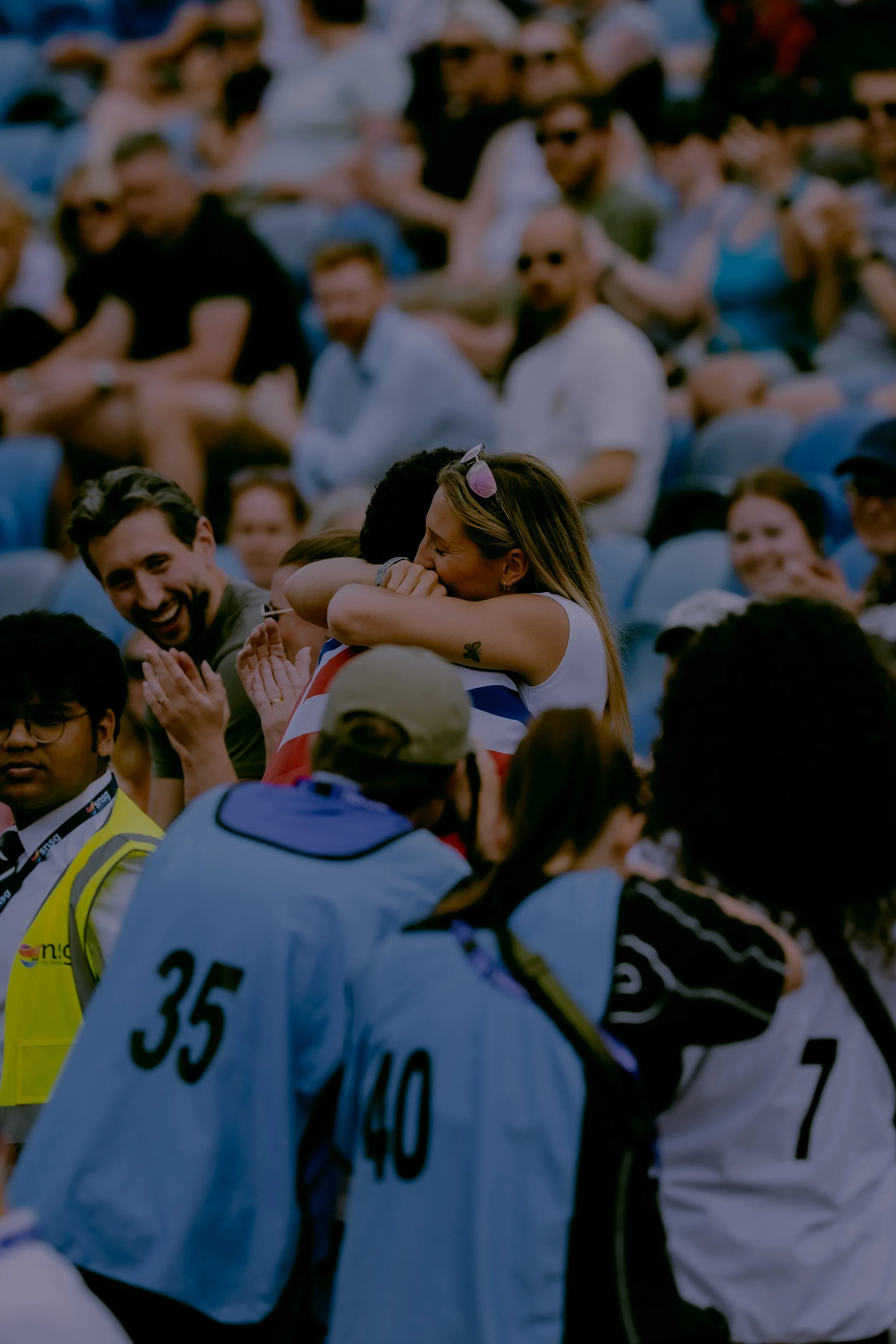A group of people gathered outdoors, some wearing sunglasses and athletic clothing, with a woman emotional and possibly crying, hugging someone. There are others smiling and interacting around them.