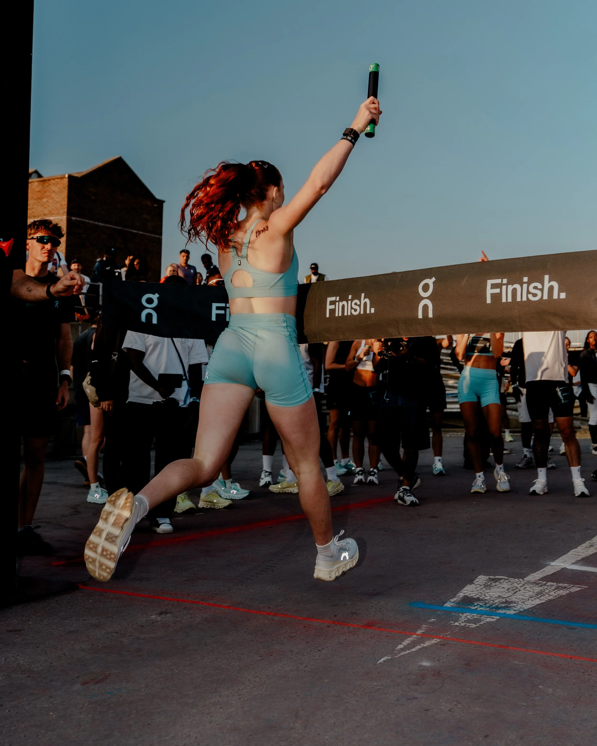 A woman with red hair celebrating after finishing a race, holding a black and green object in the air, crossing a finish line with a crowd of people in the background.