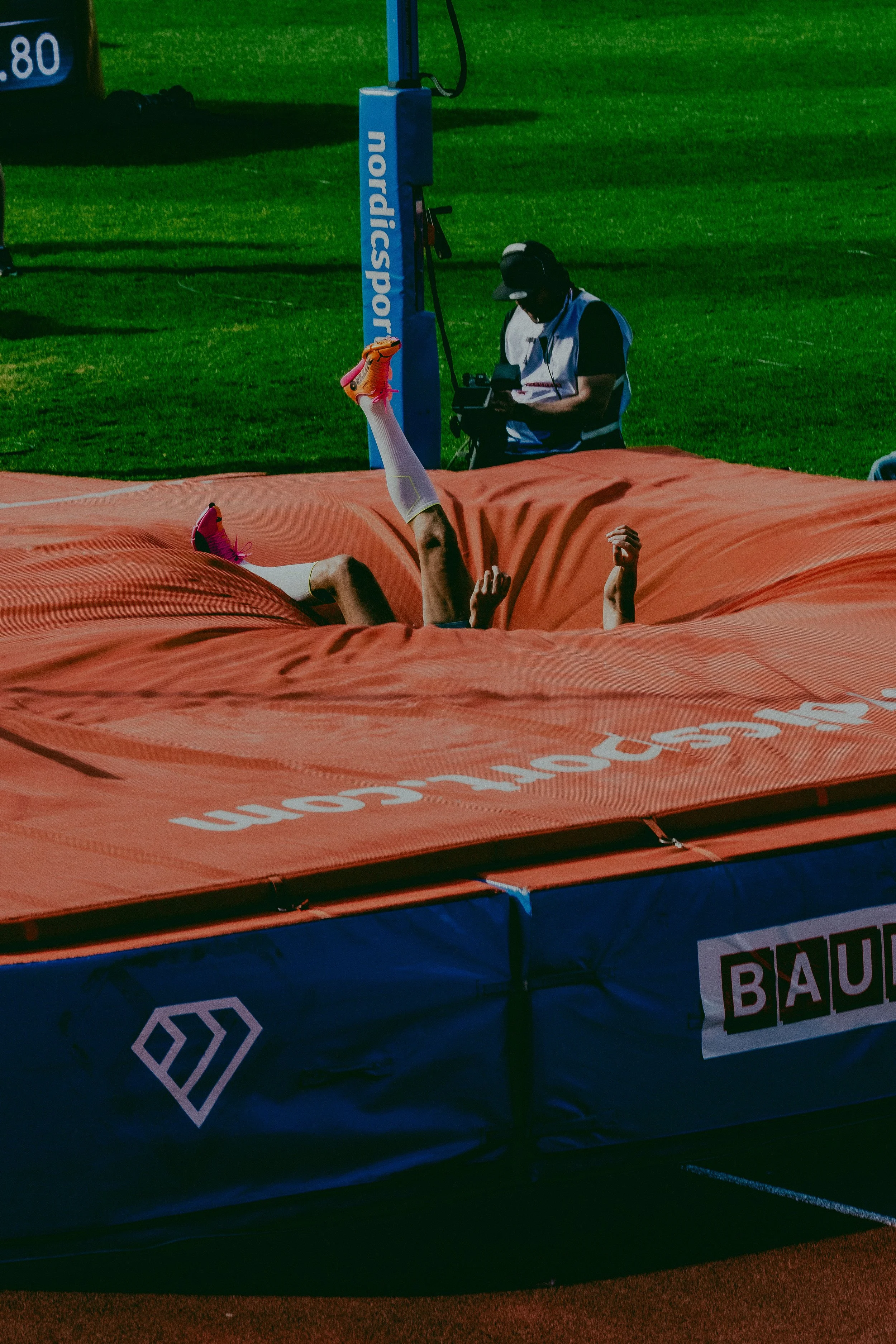 An athlete lying on an orange cushioned mat on a sports field, with legs raised and hands clenched in fists. A person in a white vest and black cap is observing and using a device nearby.