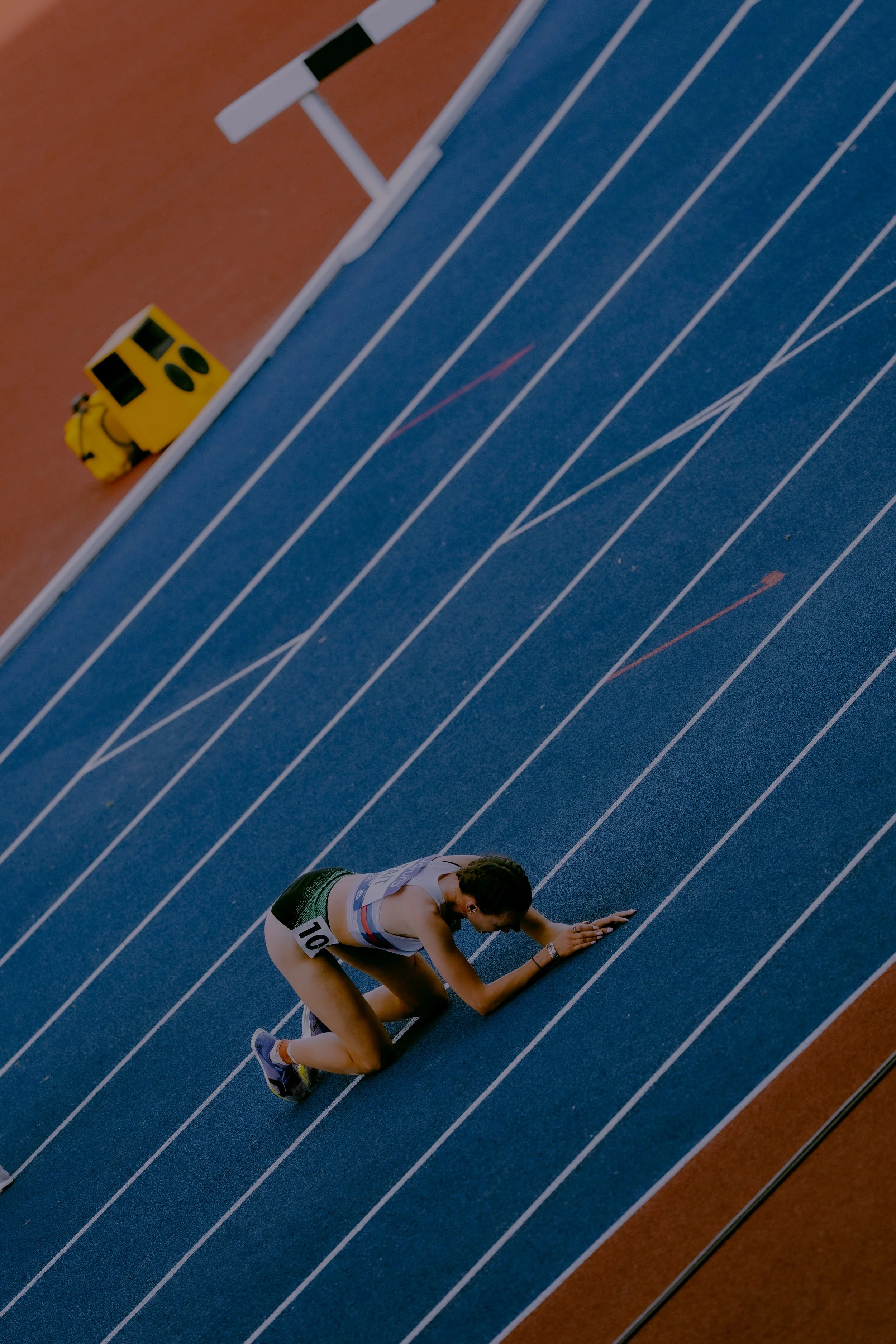 An athlete wearing a black and white uniform with the number 704 crouches on a blue running track, preparing for a race, with a yellow hurdle nearby.