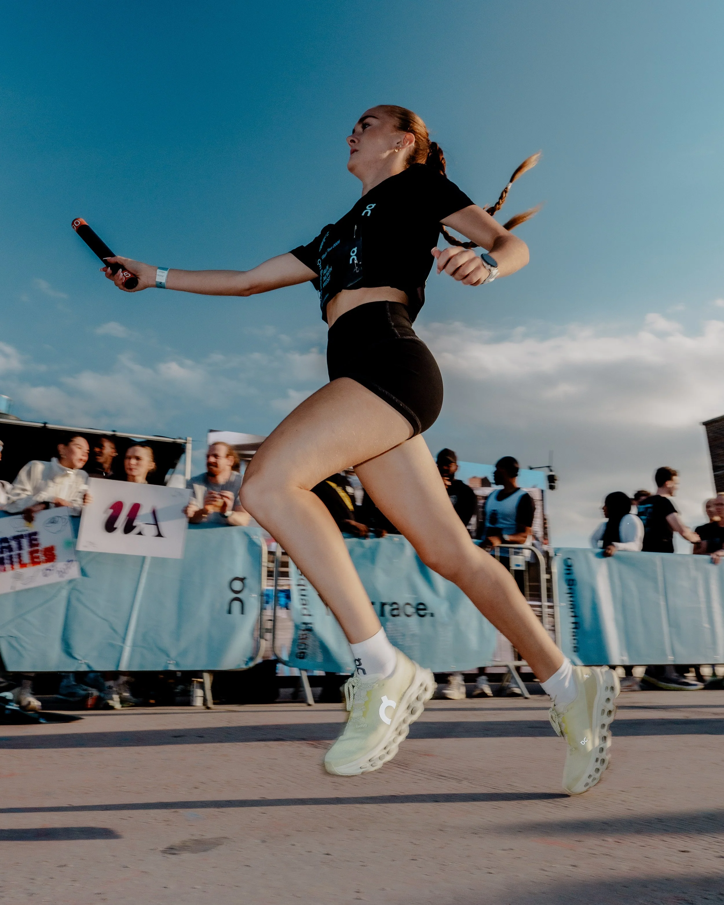 A female runner in athletic attire, wearing a black T-shirt and shorts with white sneakers, running on a track during a sports event with spectators in the background, under a clear blue sky.