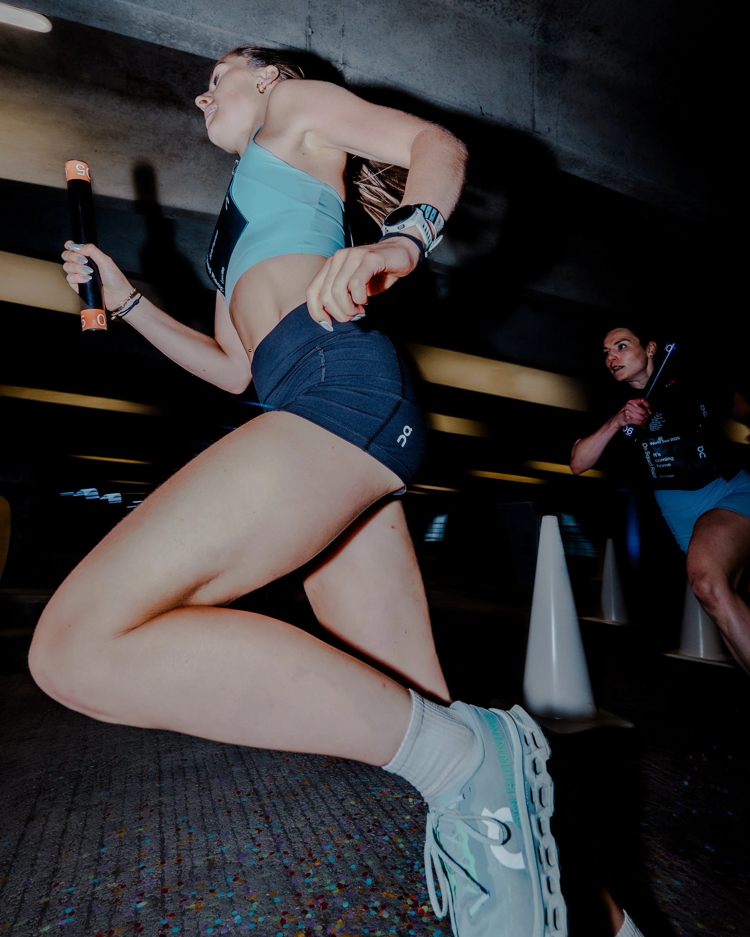 A woman running in athletic gear, wearing a light blue sports top and dark shorts, holding a baton, with a woman in the background and colorful confetti on the floor.
