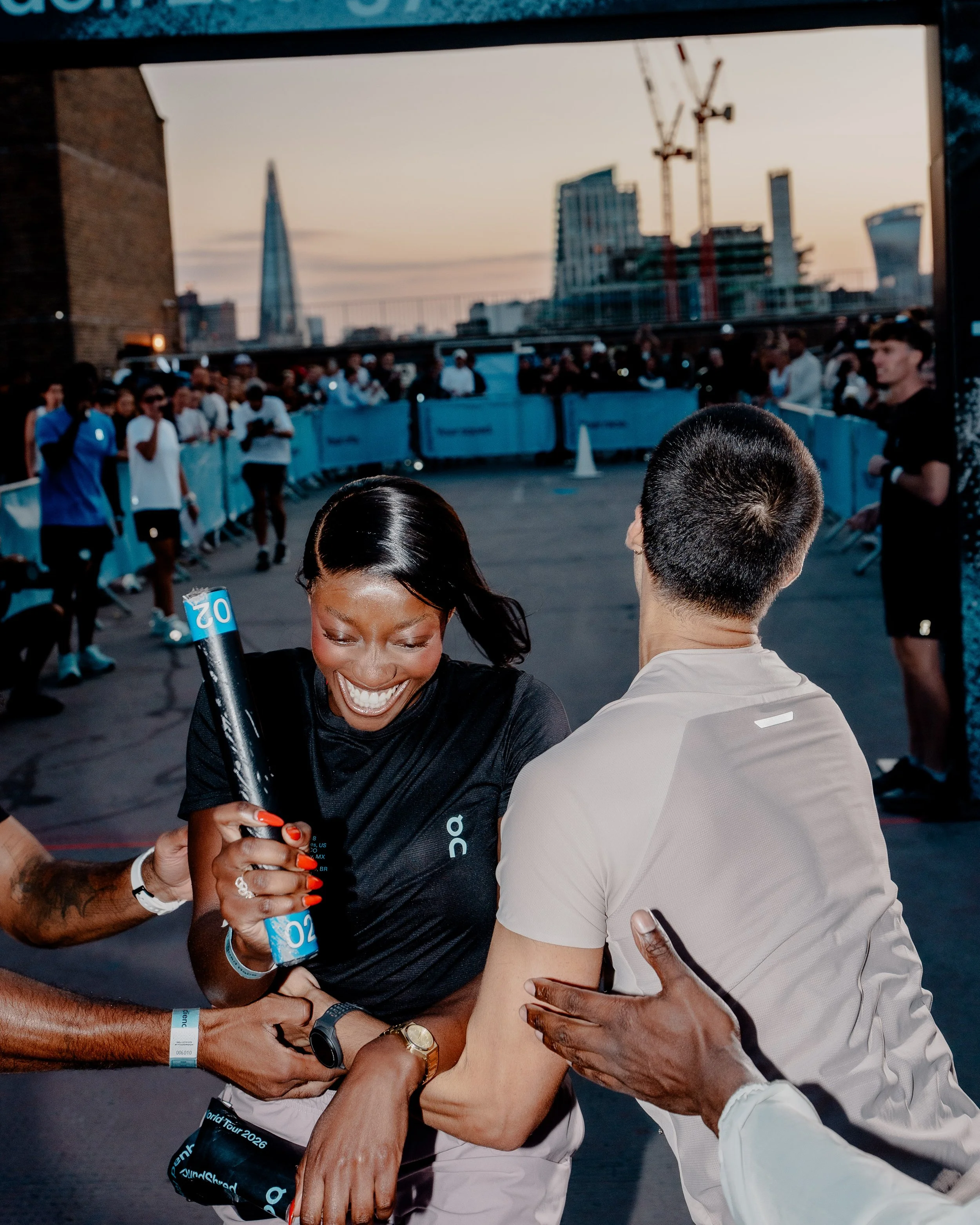 People celebrating at a marathon event, with London skyline and construction cranes in the background, as two individuals exchange friendship bracelets.