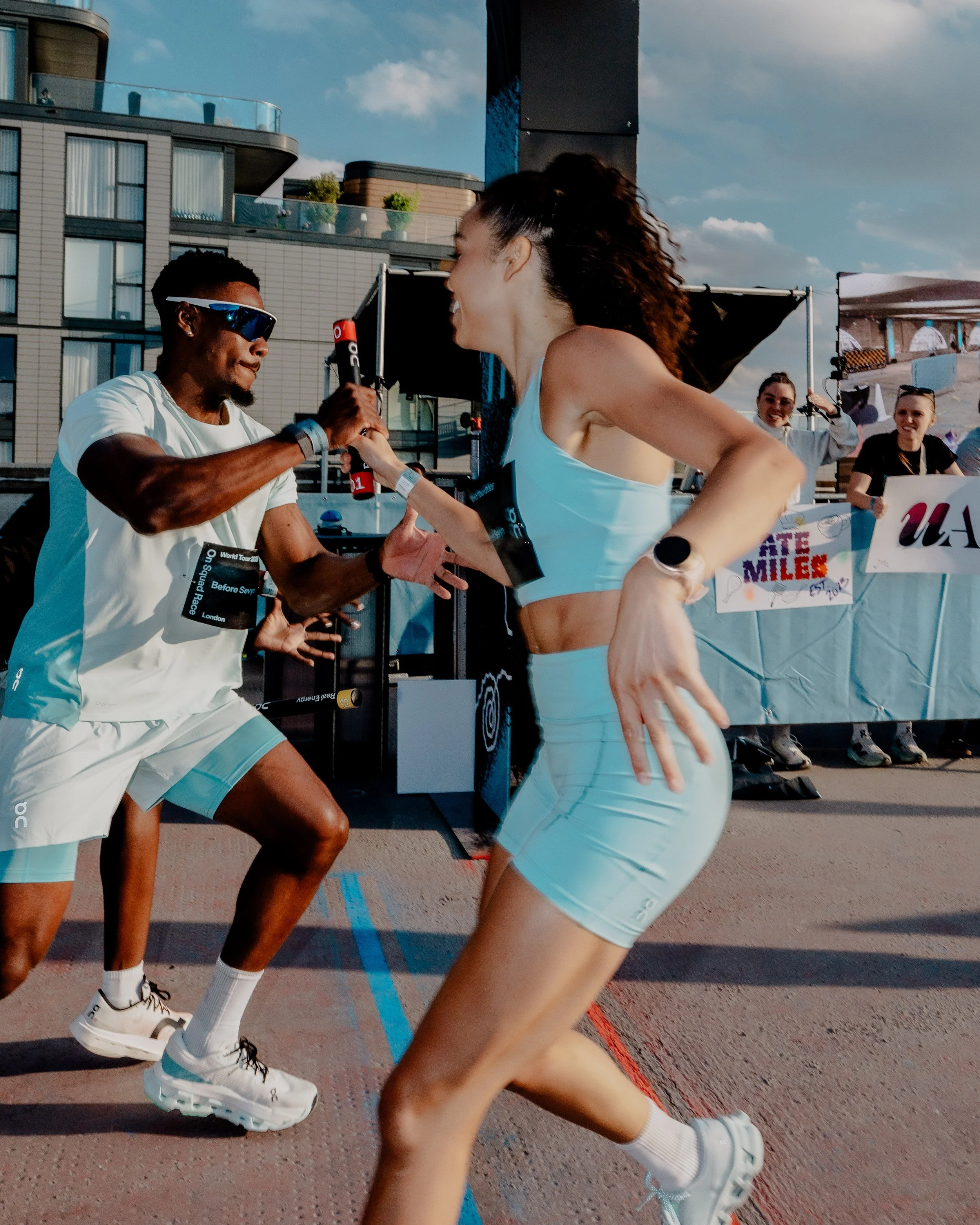 Two runners in matching light blue athletic gear passing a baton during a relay race, with a relay exchange happening on a track, and people in the background cheering, during an outdoor event in a city setting.