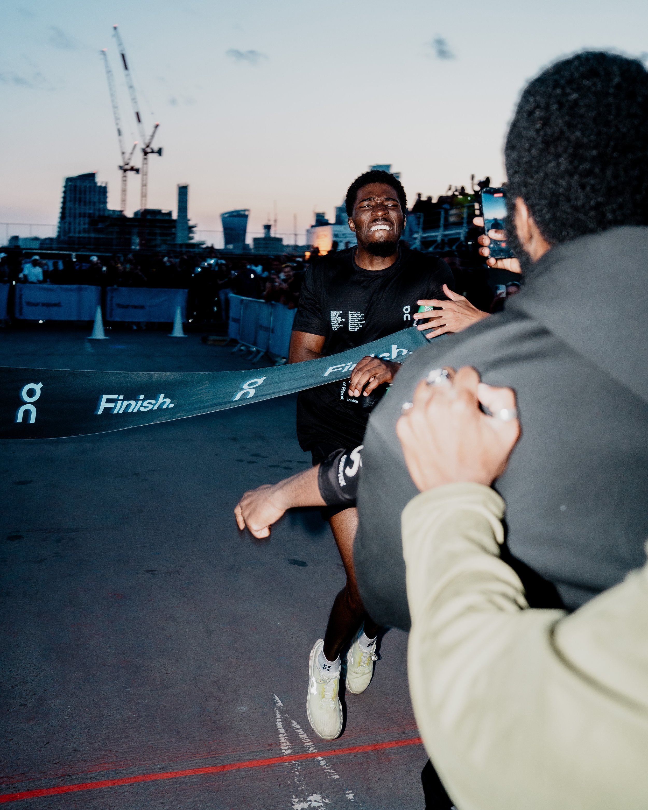 A man crosses a finish line at a race, grimacing with effort, at dusk with a city skyline in the background, as a person in the foreground holds a smartphone recording.