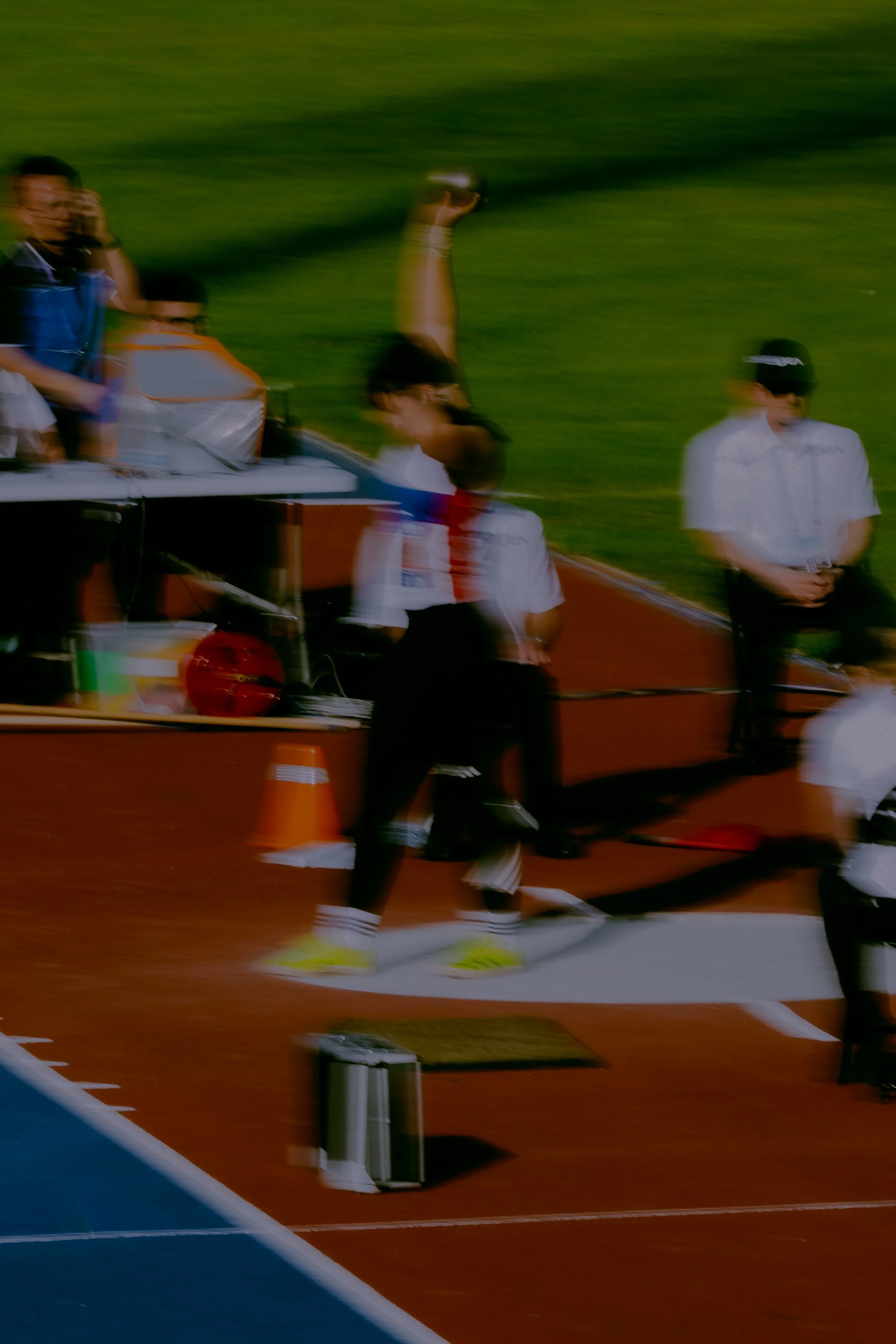 Blurred image of athletes on a red running track at an outdoor sports event, with some individuals standing and others in motion.