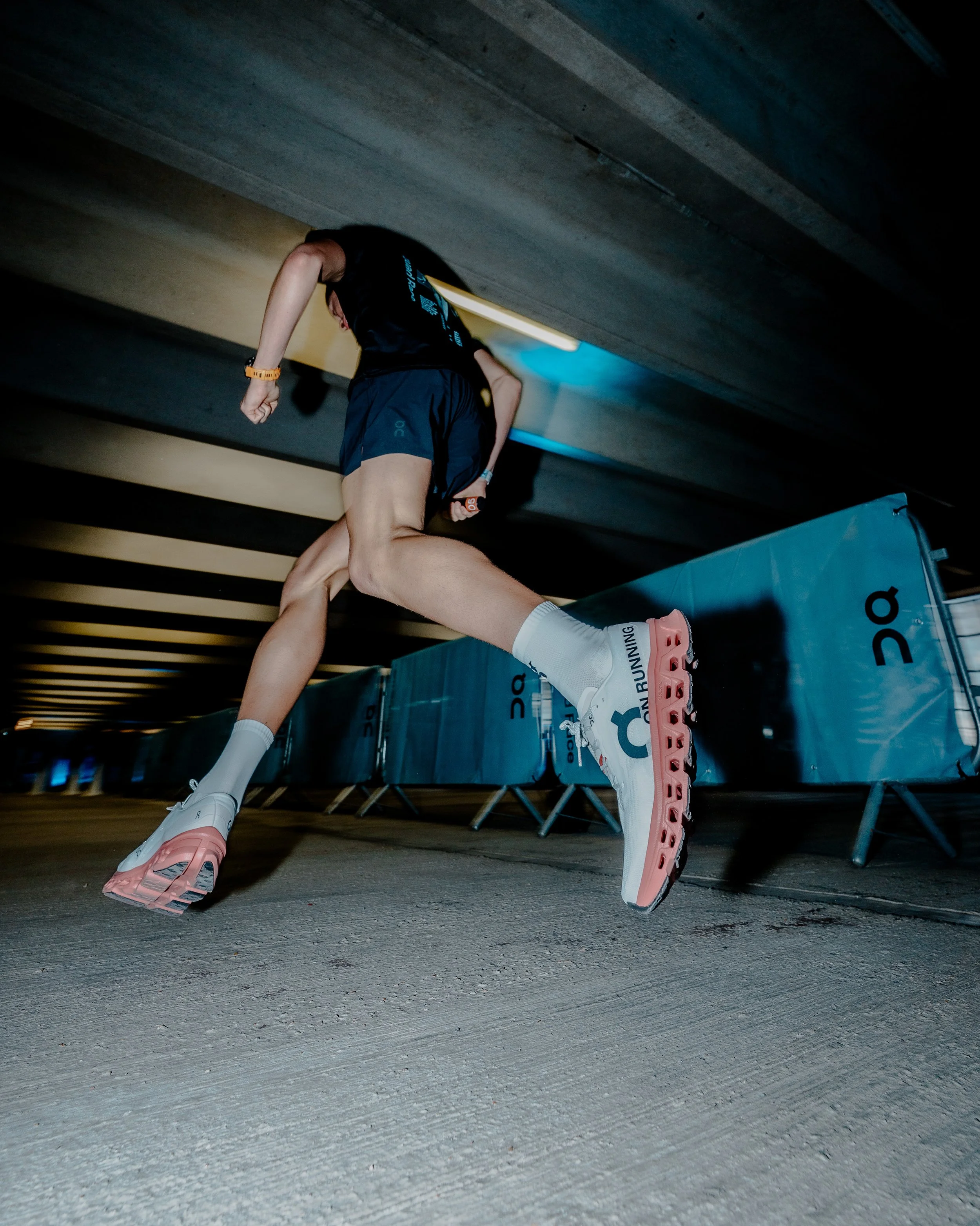 Close-up of a runner's legs and feet, wearing white and pink running shoes, on a concrete floor, with a background of wooden ceiling beams.