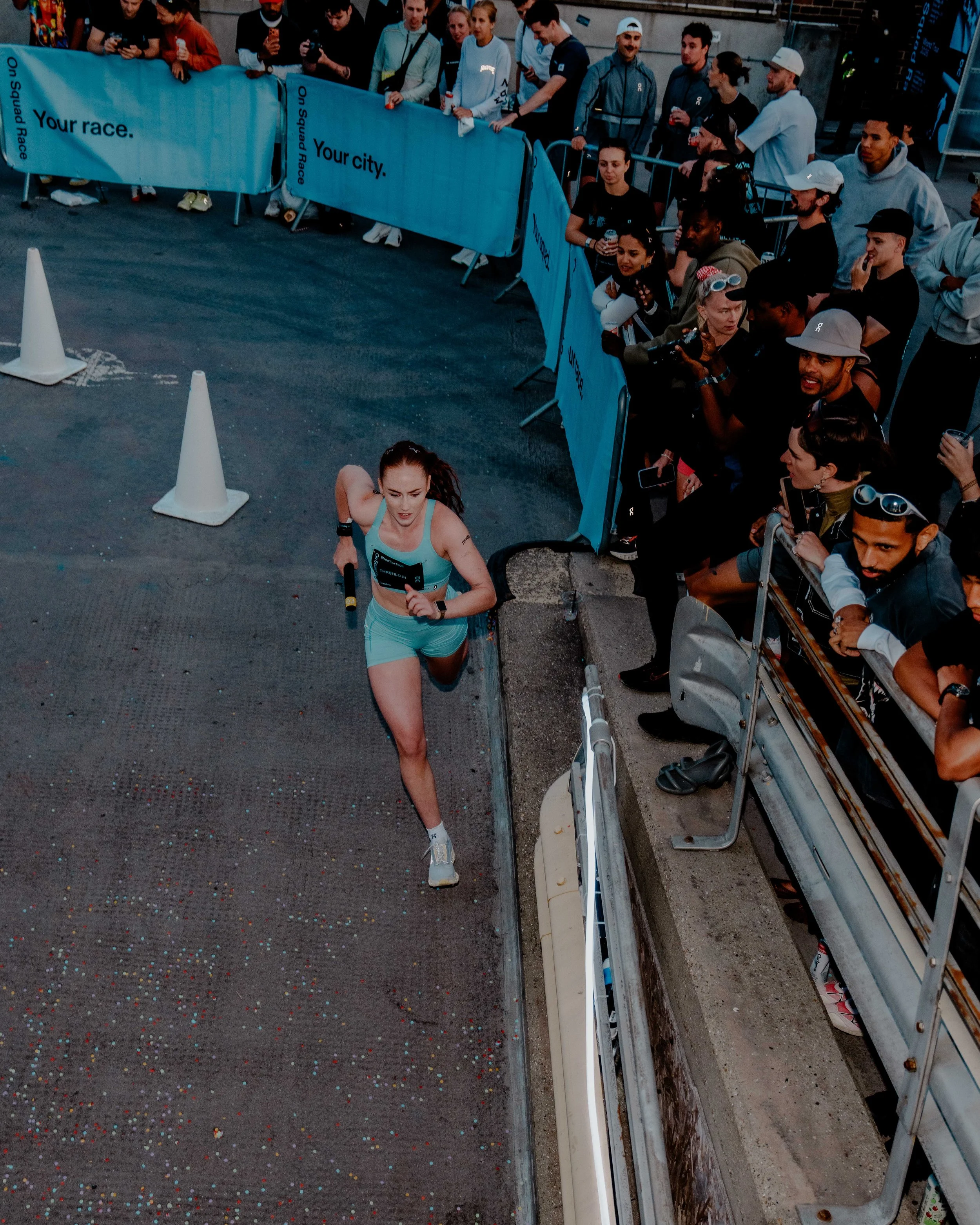 A woman running in a race with a crowd of spectators watching along the side of the course, which is marked by police barriers and traffic cones.