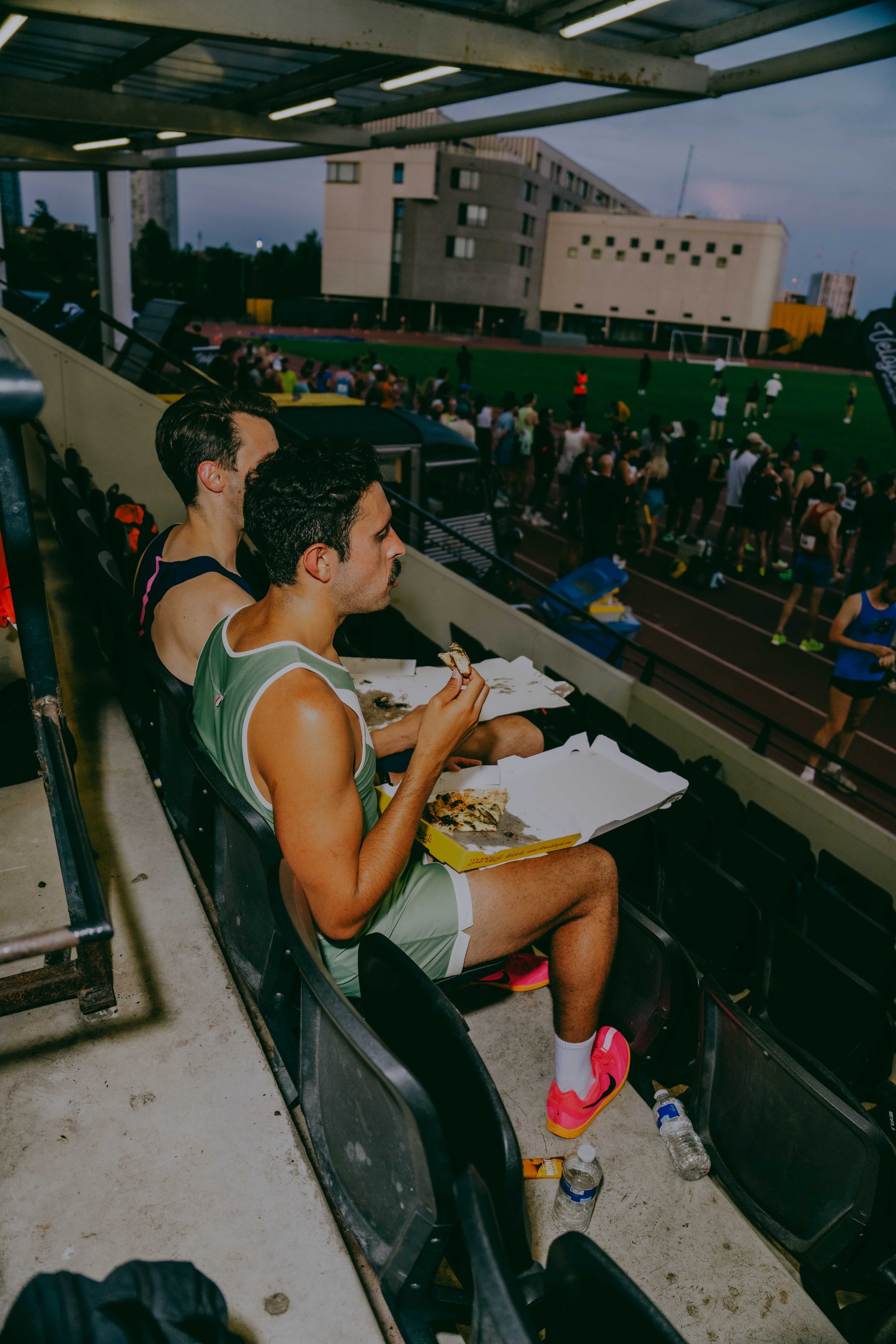 Two men dining on pizza in the stands of an outdoor sports stadium during an event, with a crowd and a field visible in the background.